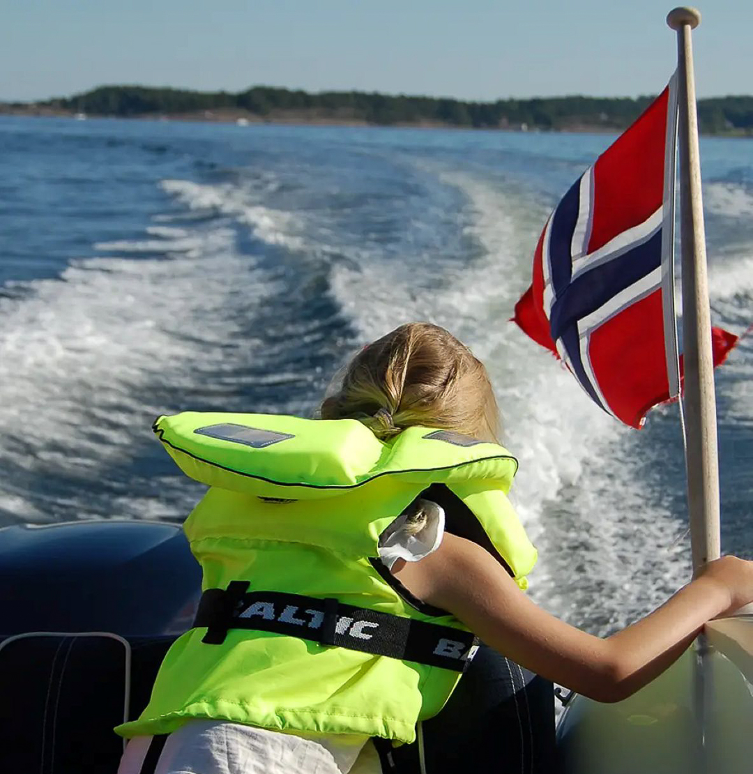 Jente med gul redningsvest sitter bakerst i båt i fart og ser på kjølvannet fra båten.