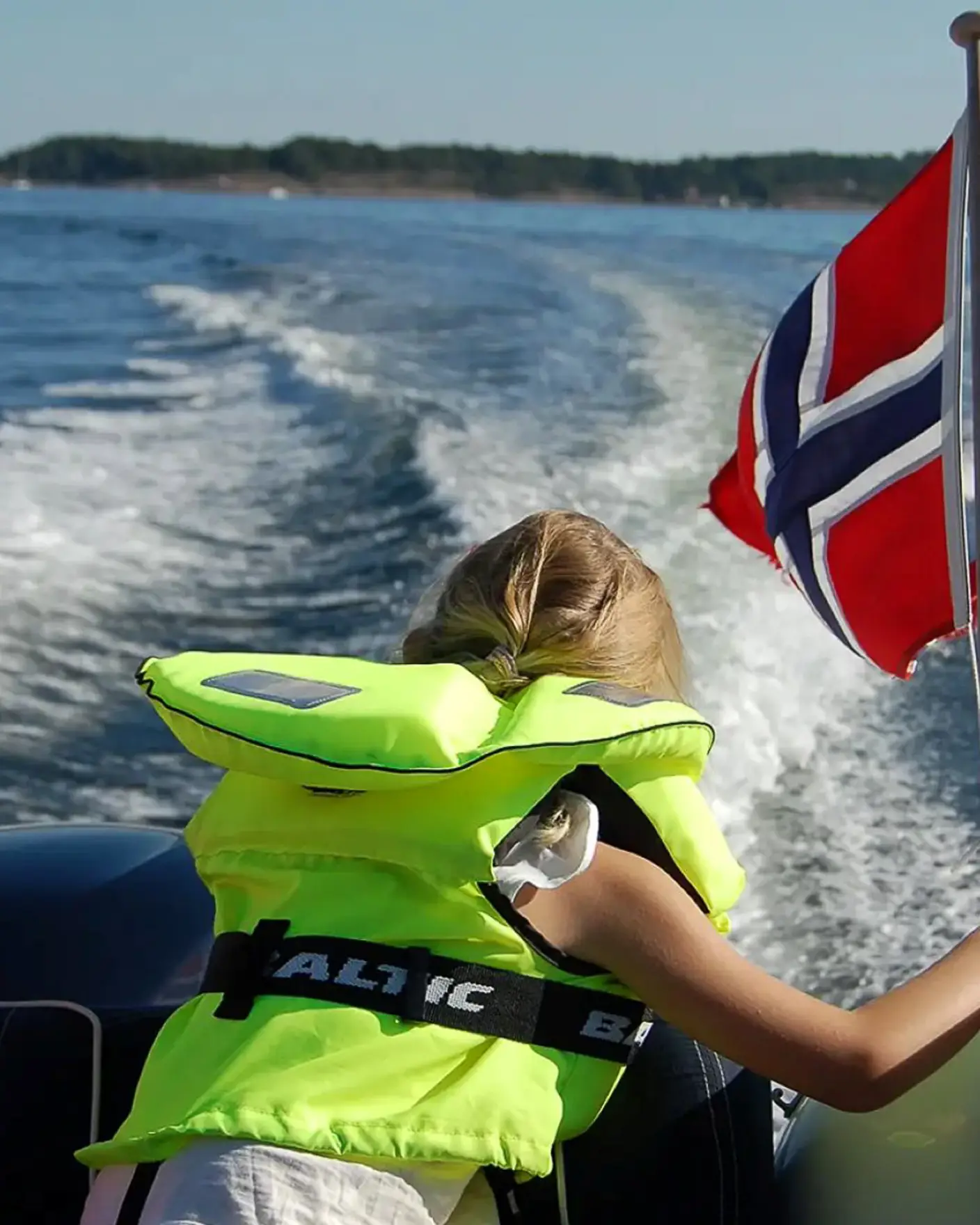 Jente med gul redningsvest sitter bakerst i båt i fart og ser på kjølvannet fra båten.