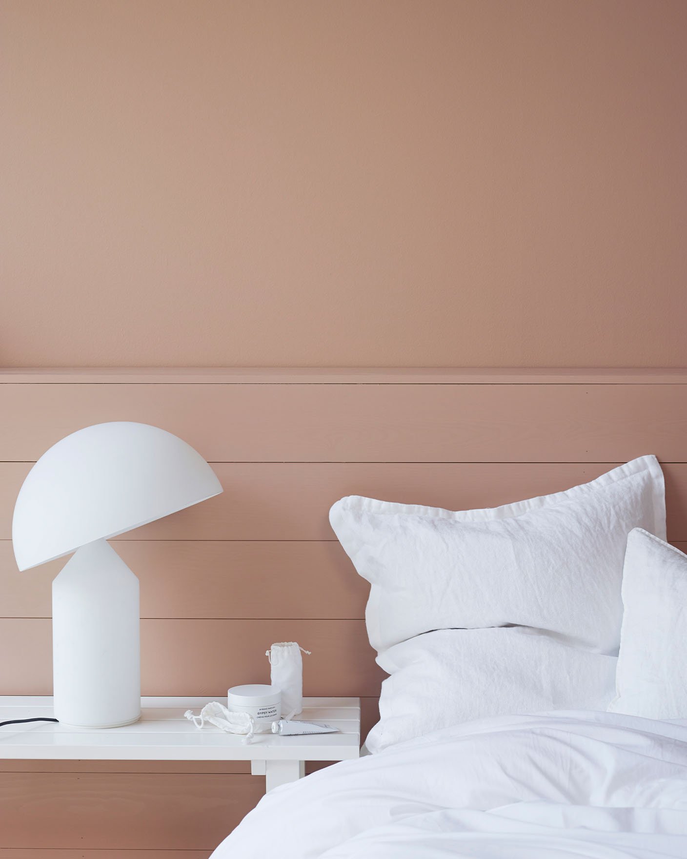 Närbild av en säng med krispigt vitt sänglinne och en skulptural vit lampa, mot en dämpad gyllenrosa vägg målad med Jotun 2024 Senses.