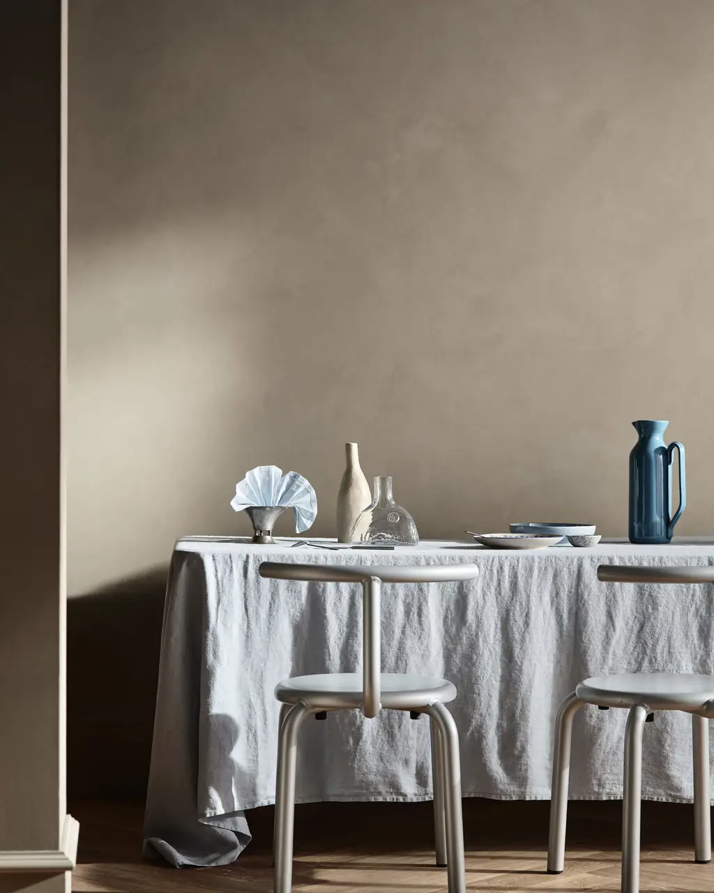 A dining table set with blue and clear glass pitchers before textured walls in Jotun 10966 Almond Beige Touch of Suede