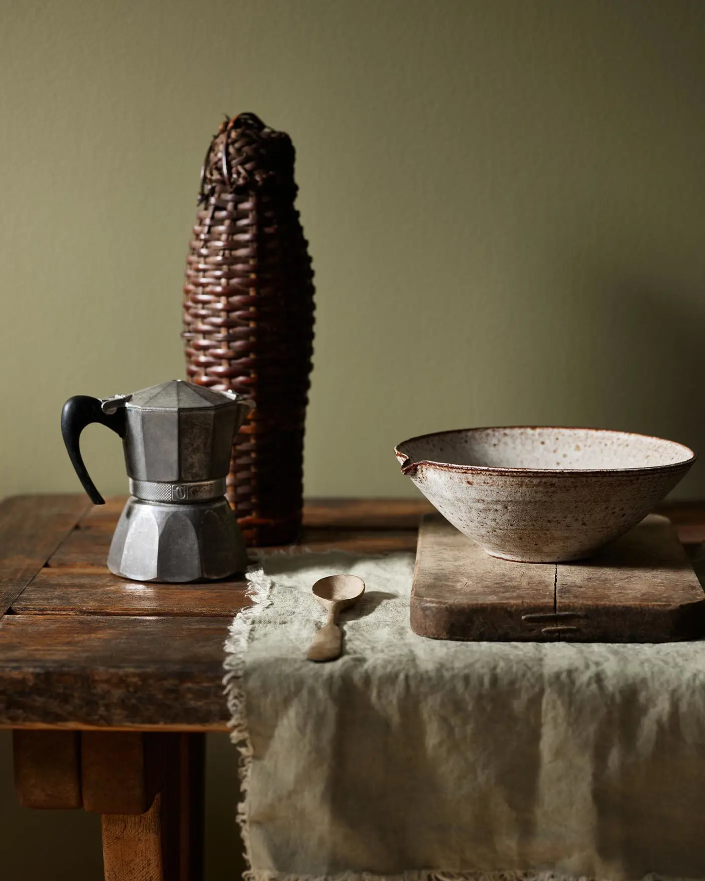 Rustic wooden table with a metal espresso maker and ceramic bowl with a olive green background painted with the Jotun colour 8597 Seaweed Green.
