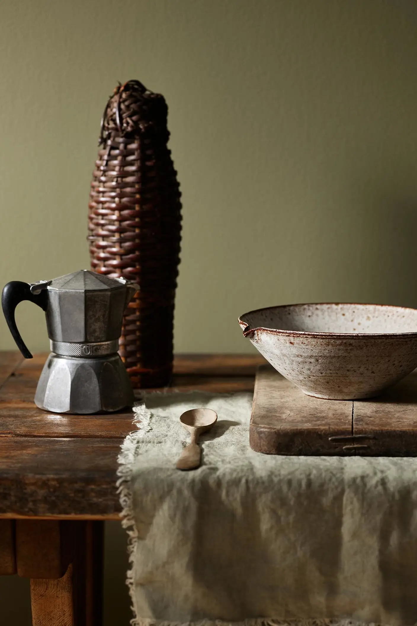 Rustic wooden table with a metal espresso maker and ceramic bowl with a olive green background painted with the Jotun colour 8597 Seaweed Green.