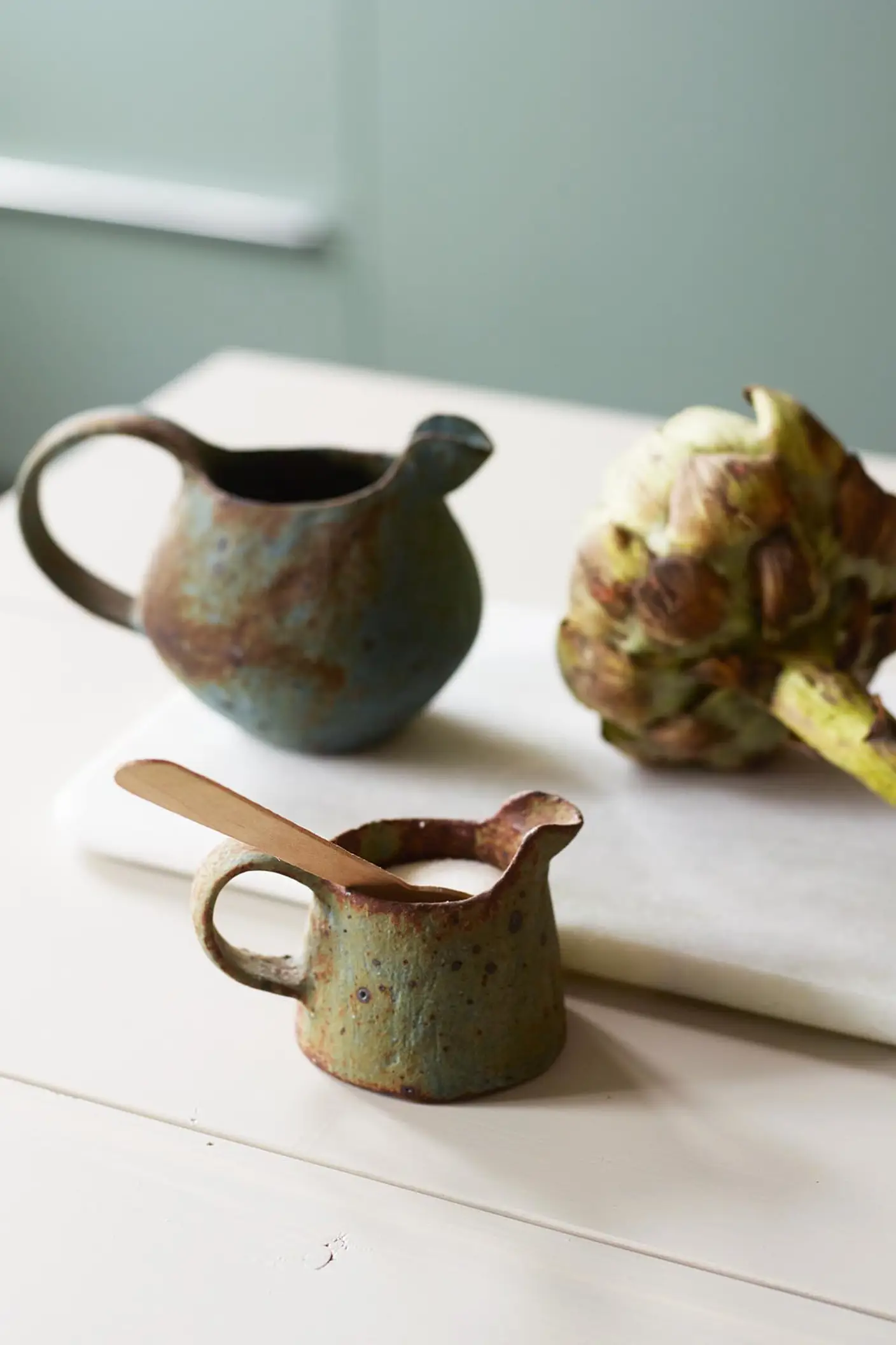 Rustik keramik och en färsk kronärtskocka vilar på en ljus yta, medan mjuka gröna väggar målade med Jotun 7163 Minty Breeze skapar en sval och uppfriskande bakgrund i rummet.