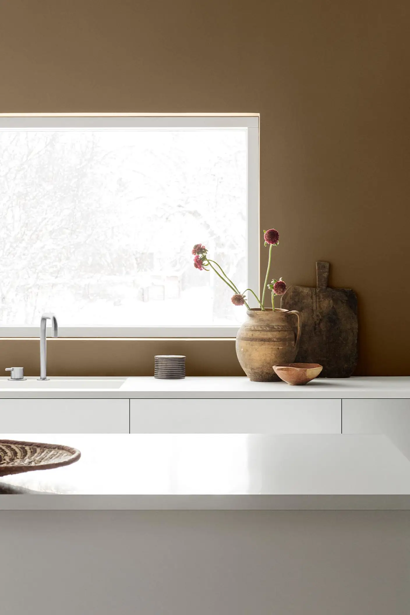 Beyaz dolaplar ve sade dekorlarla düzenlenen minimalist mutfak; Jotun 10963 Golden Bronze ile boyanan altın kahverengi duvarın önünde sıcak bir görünüm sunuyor.