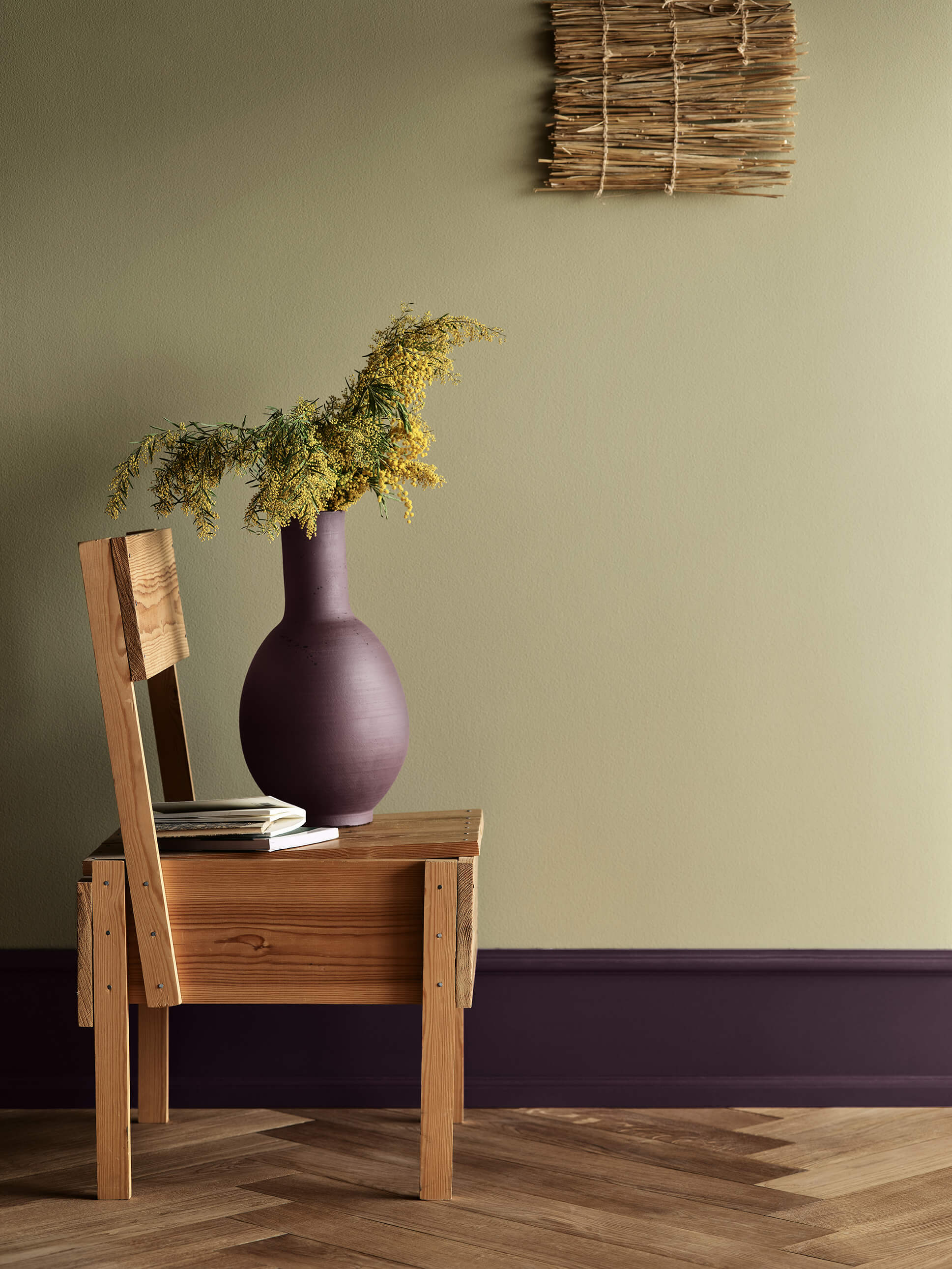 En stue med en vegg i gulgrønn med Jotun 0290 Biri og gulvlister i dyp lilla med Jotun 3570 Velvet Plum, med en trestol hvor det står en lilla blomstervase