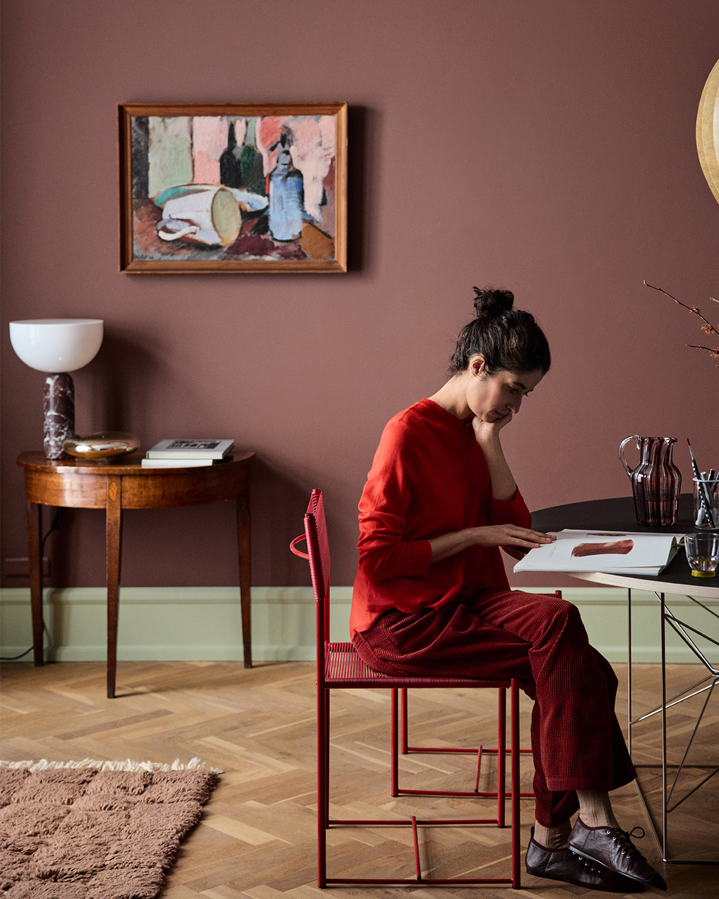 Jotun 2951 Sophisticated Red ile boyanmış koyu kırmızı duvarlı modern odada, kırmızı kıyafetli bir kadın kırmızı sandalyede oturup masada kitap inceliyor; duvarda sanat eseri yer alıyor.