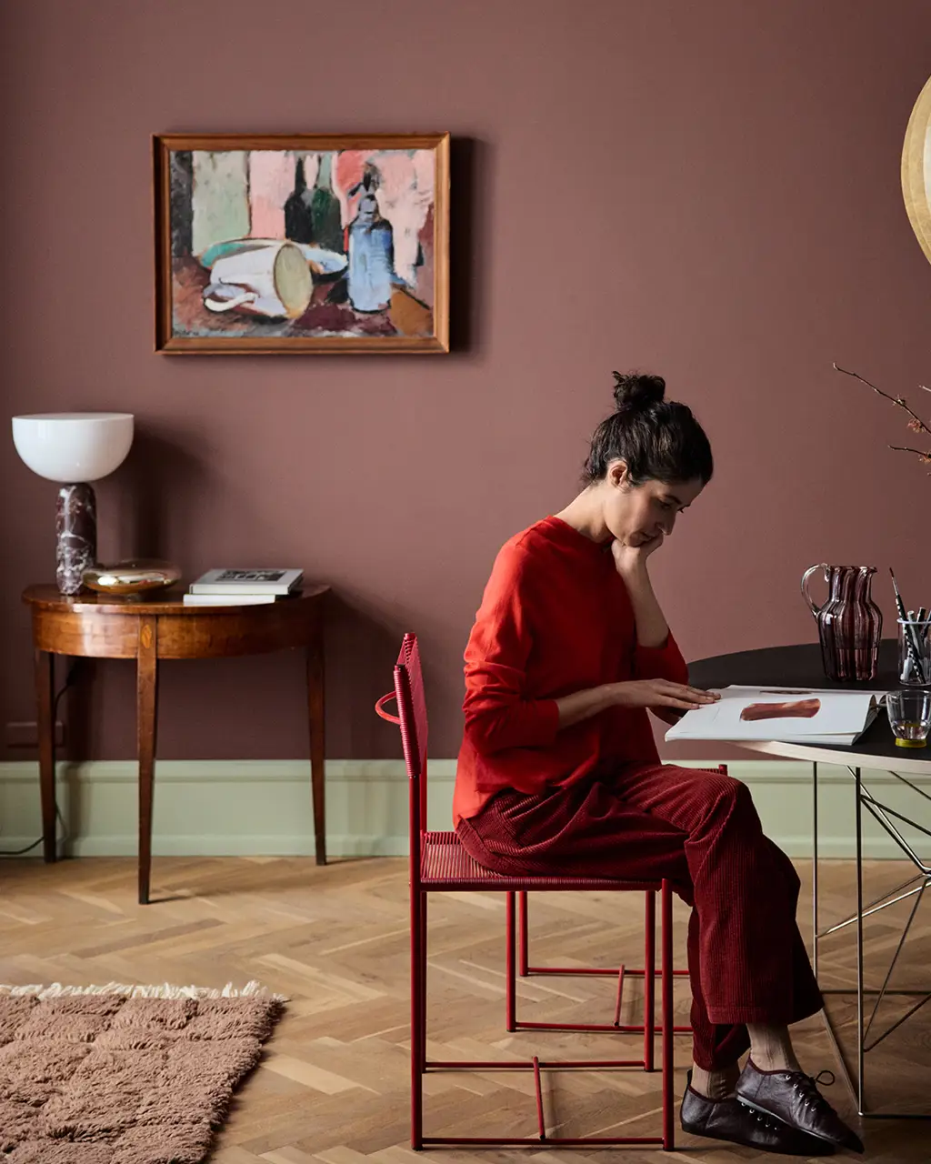 Jotun 2951 Sophisticated Red ile boyanmış koyu kırmızı duvarlı modern odada, kırmızı kıyafetli bir kadın kırmızı sandalyede oturup masada kitap inceliyor; duvarda sanat eseri yer alıyor.
