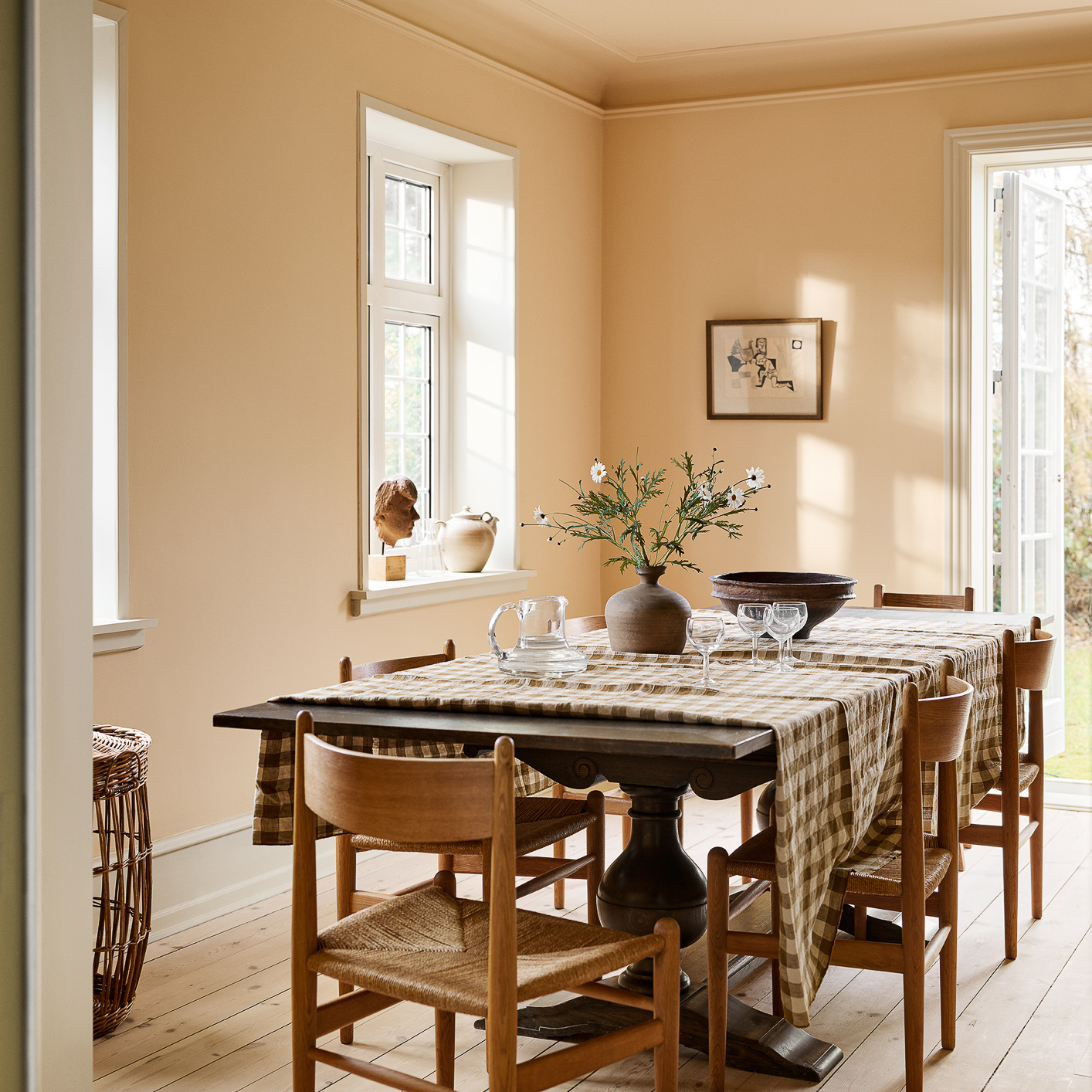 Spisestue med møbler i mørkt tre og bord med brunrutet duk mot vegger malt i gyllen okergul, Jotun Mild Ochre og listverk i lys gulbeige Jotun Soft Radiance