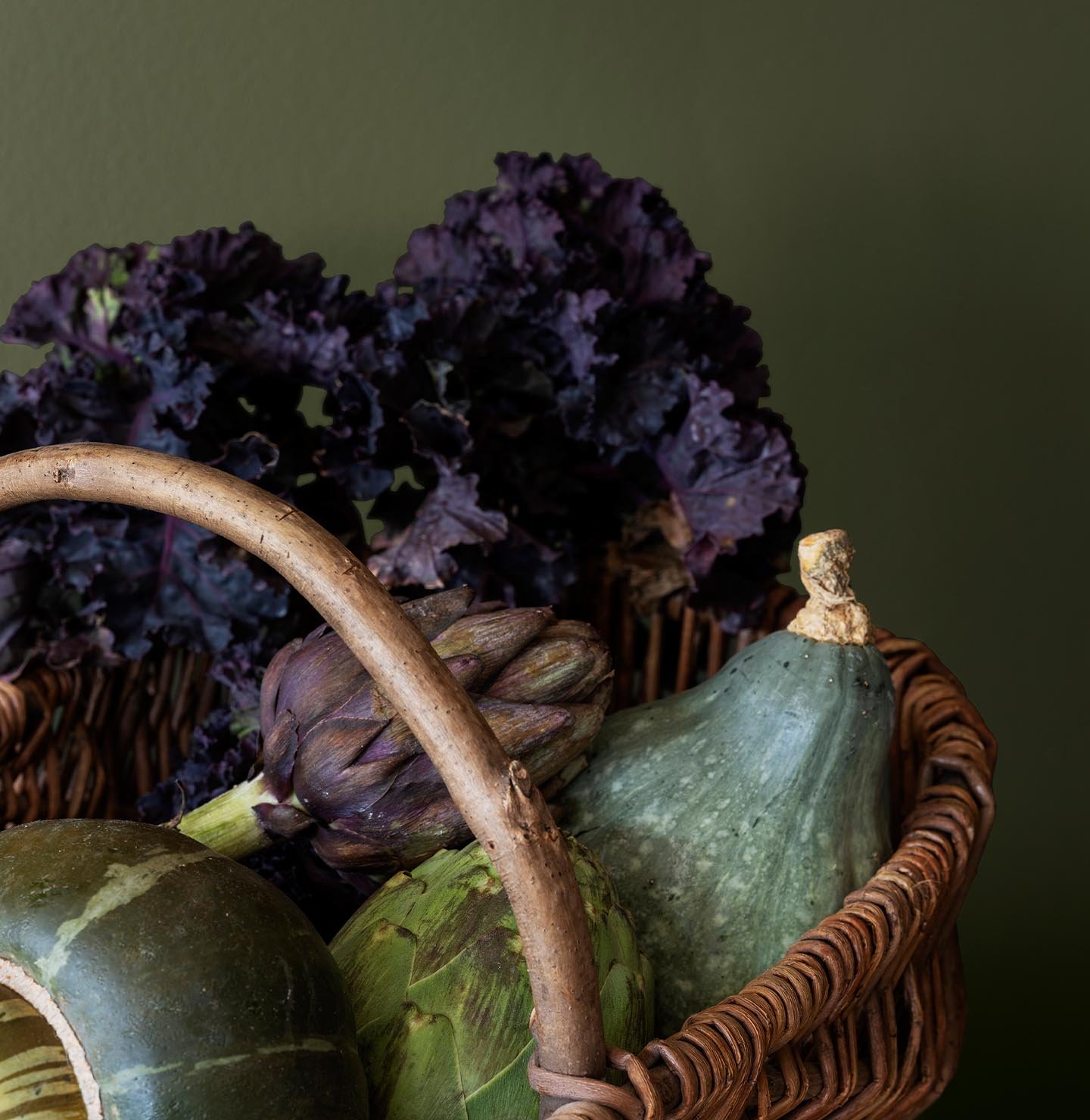 A basket filled with fresh vegetables like artichokes and kale, placed against a rich, yellowish green wall painted with Jotun 8575 Natural Green.