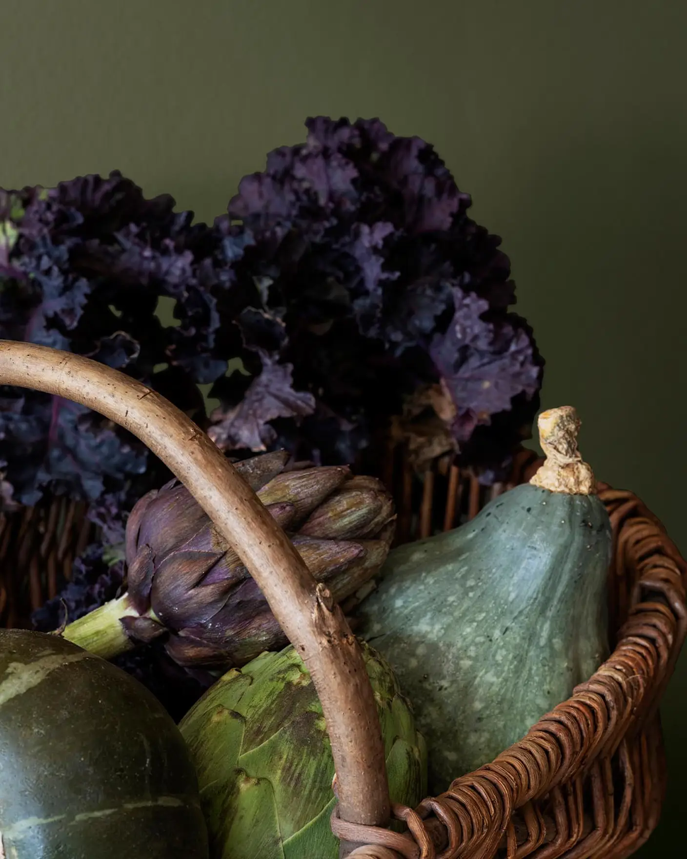 A basket filled with fresh vegetables like artichokes and kale, placed against a rich, yellowish green wall painted with Jotun 8575 Natural Green.