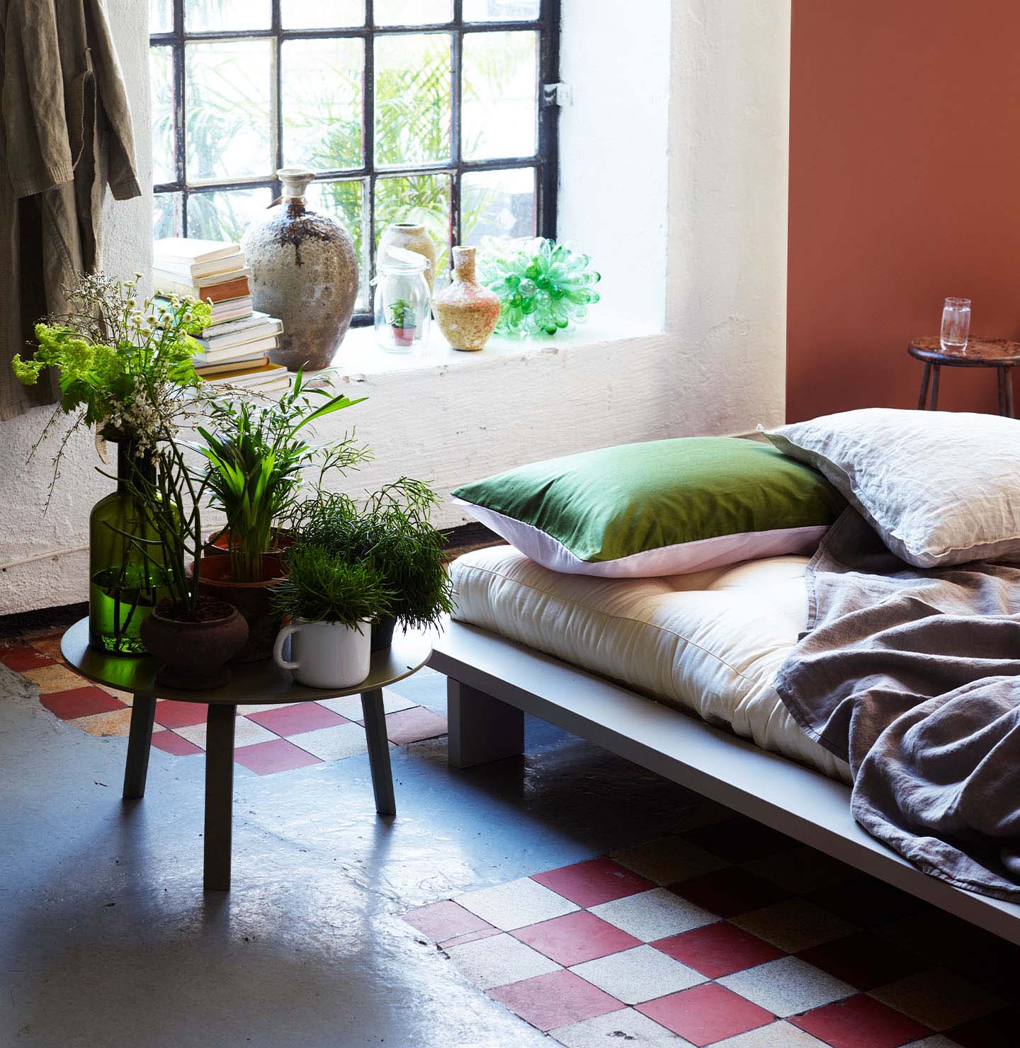 Eklektiskt sovrum med en säng klädd i neutrala och gröna textilier, omgiven av krukväxter, bredvid ett stort fönster i industristil och en terrakottafärgad accentvägg målad med Jotun 2771 Rustik Terracotta.