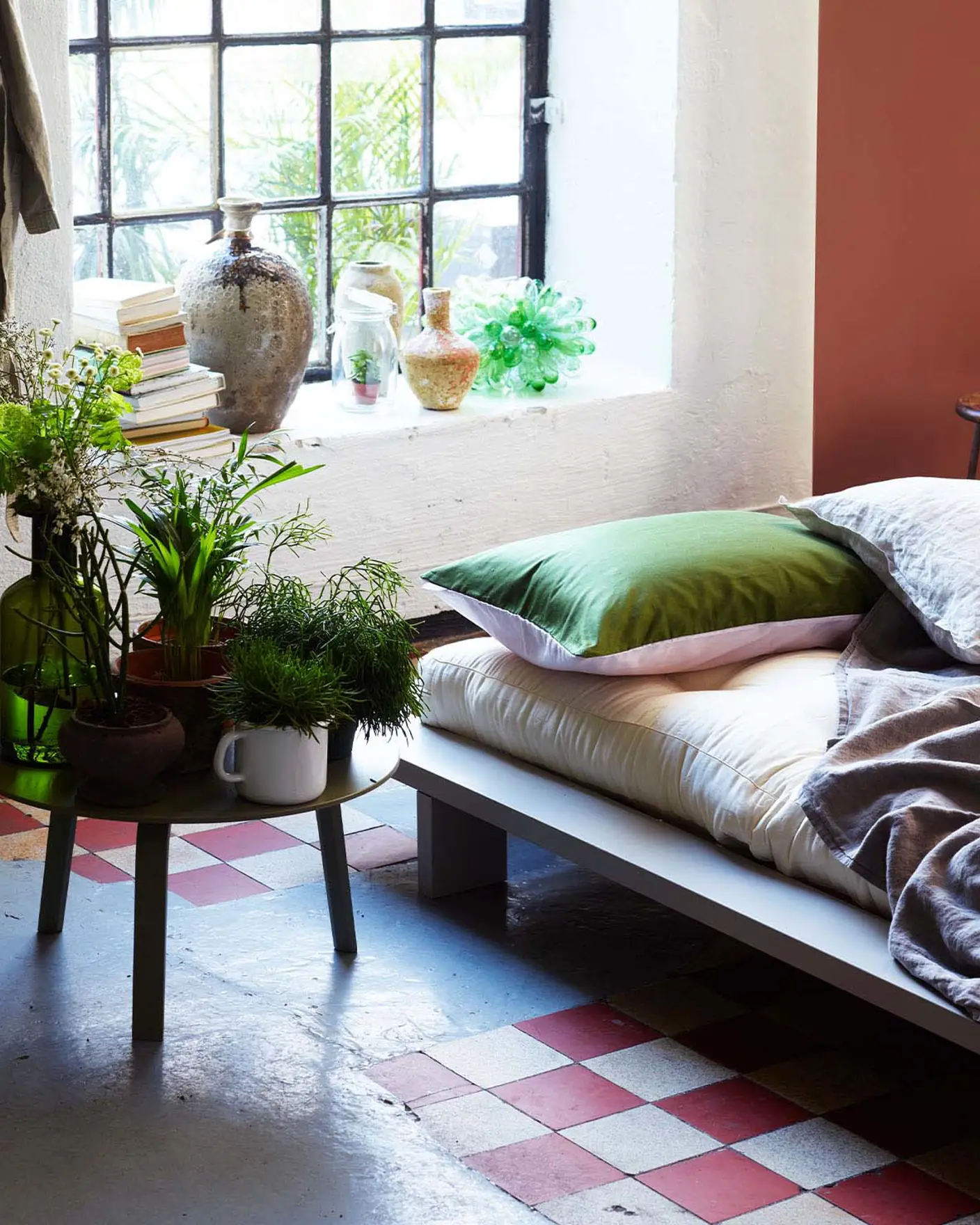 Eklektiskt sovrum med en säng klädd i neutrala och gröna textilier, omgiven av krukväxter, bredvid ett stort fönster i industristil och en terrakottafärgad accentvägg målad med Jotun 2771 Rustik Terracotta.