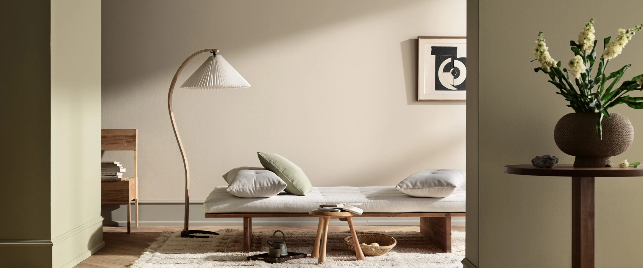 Ljust och stilrent sovrum med låg bäddsoffa, golvlampa, dekorativa kuddar och vas med blommor i nordisk inredningsstil