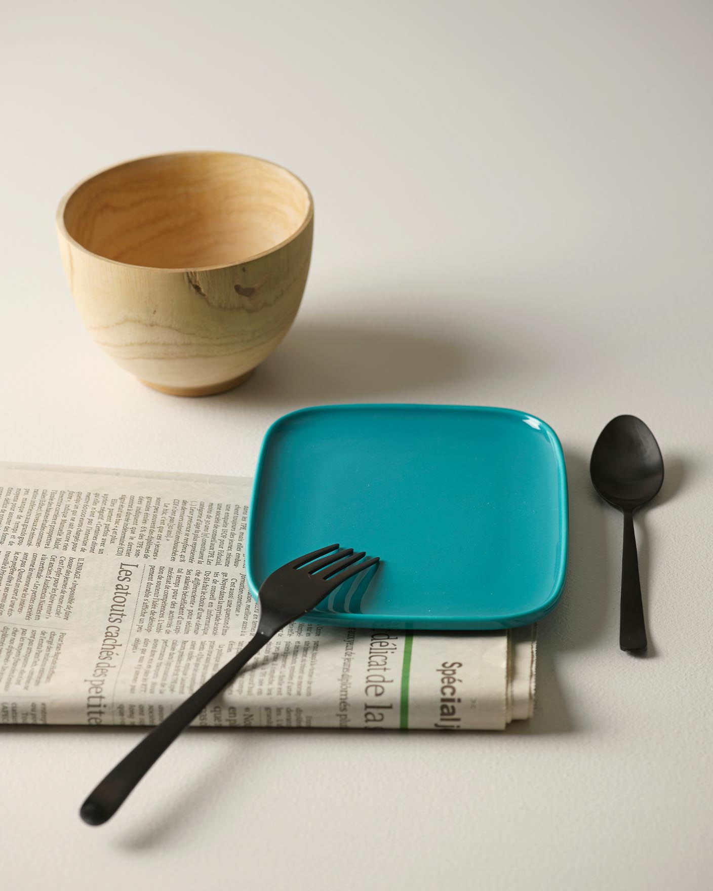 Ett bord med en teal-färgad tallrik, svart bestick, en träskål och en tidning på en ljus, gyllenvit bakgrund målad med Jotun 1624 Lätthet.