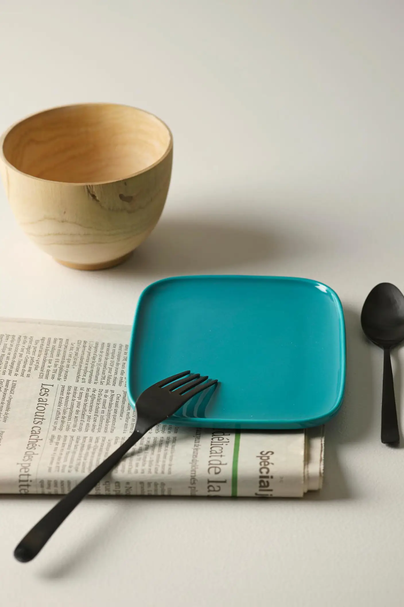 Una mesa con un plato color teal, cubiertos negros, un cuenco de madera y un periódico sobre un fondo blanco dorado claro pintado con Jotun 1624 Skylight.