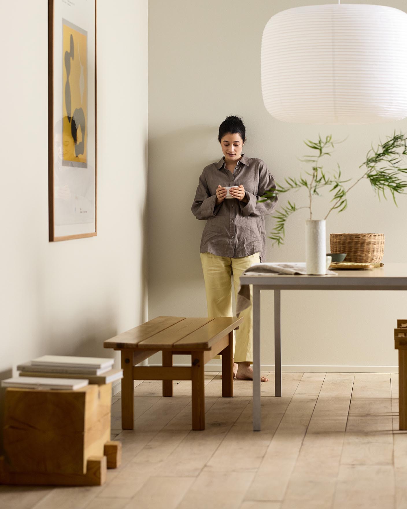 Woman in a minimalist room with wooden bench, table decor, and framed artwork against a beige wall painted in the Jotun colour 12308 Unbleached.
