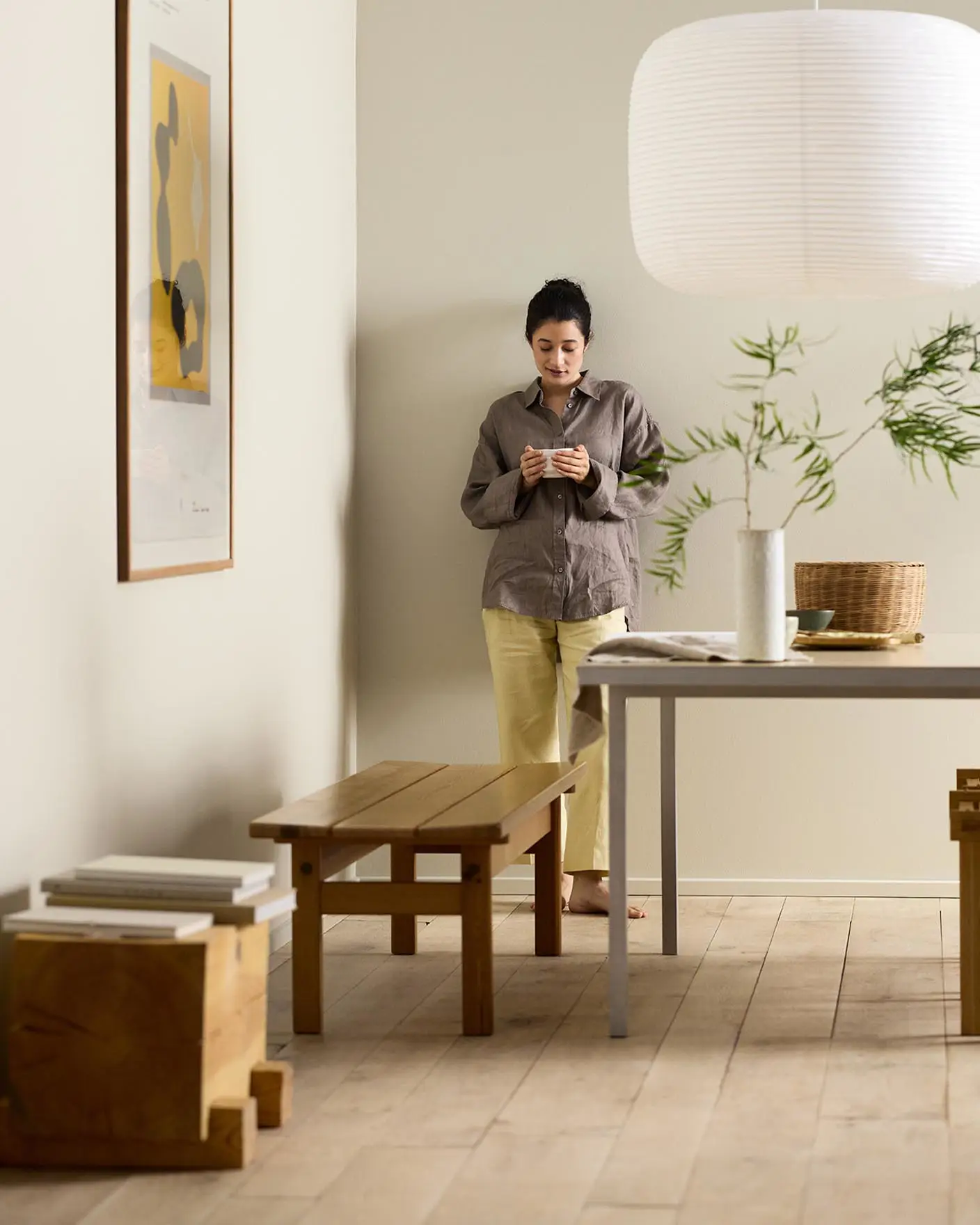 Woman in a minimalist room with wooden bench, table decor, and framed artwork against a beige wall painted in the Jotun colour 12308 Unbleached.