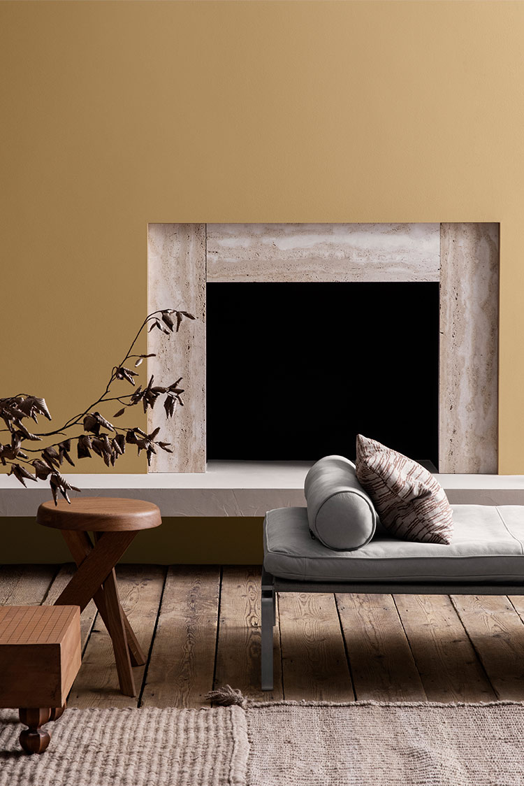 Moderne stue med minimalistisk dekor som har en okergul vegg malt i Jotun 1909 Chalk Ochre, åpen peis, tregulv og stilige møbler inkludert en brun vase, trebord og grå chaiselong