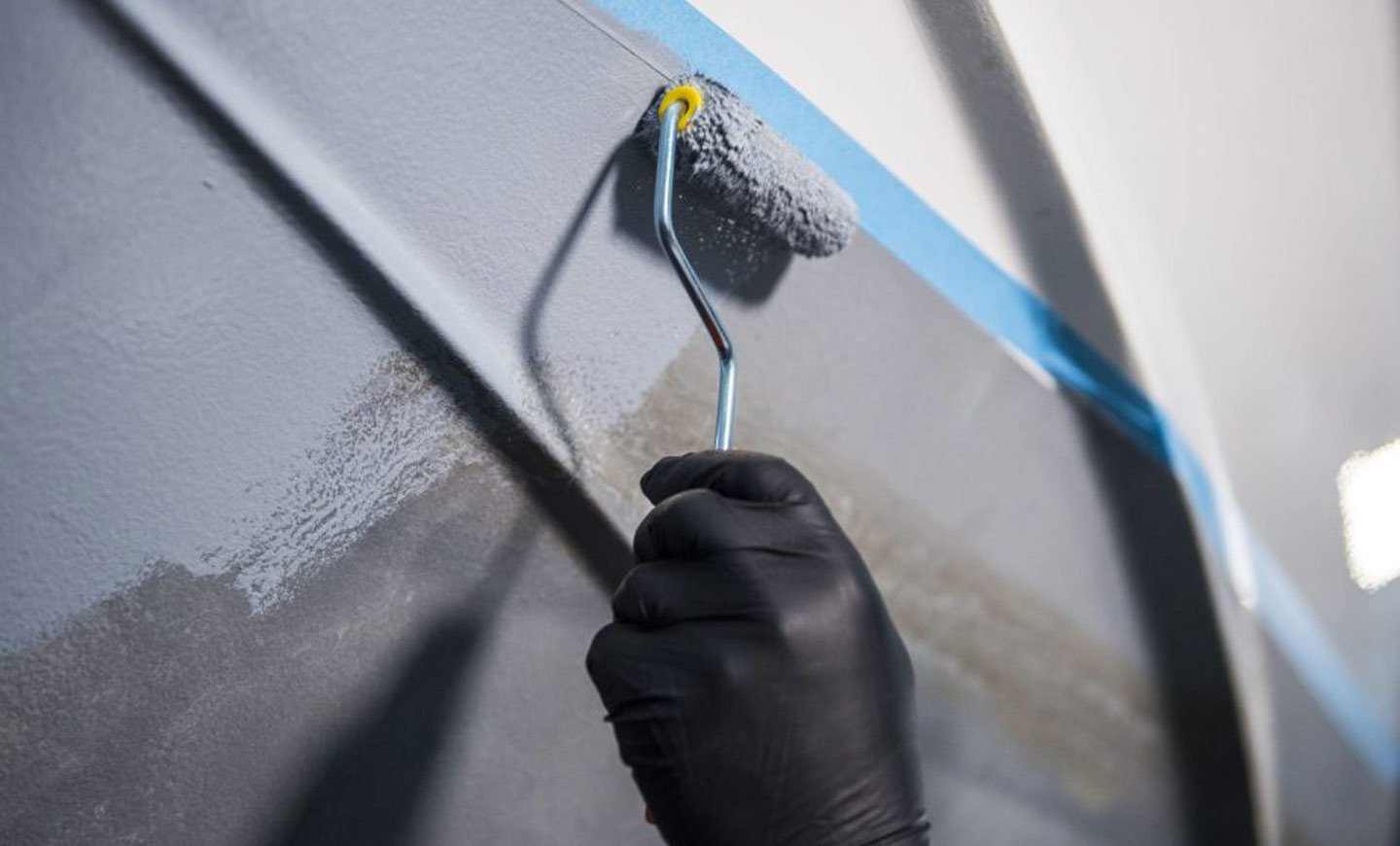 Hånd med sort hanske holder malingsrulle og påfører grunning av båt med produkter fra Jotun