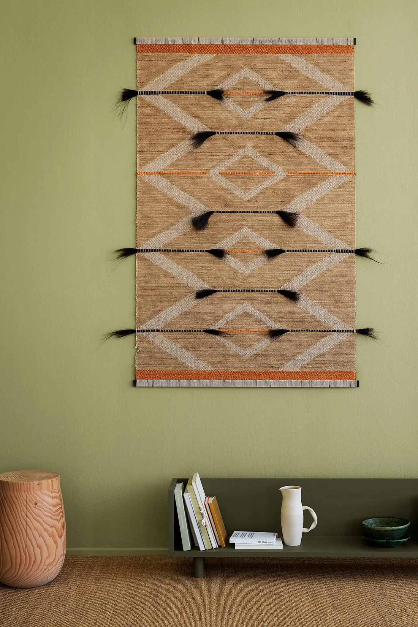 Böcker och keramik vilar på en grön hylla i färgen 8575 Natural Green från Jotun, med en vävd matta på väggen och en mjuk olivgrön bakgrund målad med Jotun 8183 Örtagård, som tillför naturlig värme till rummet.