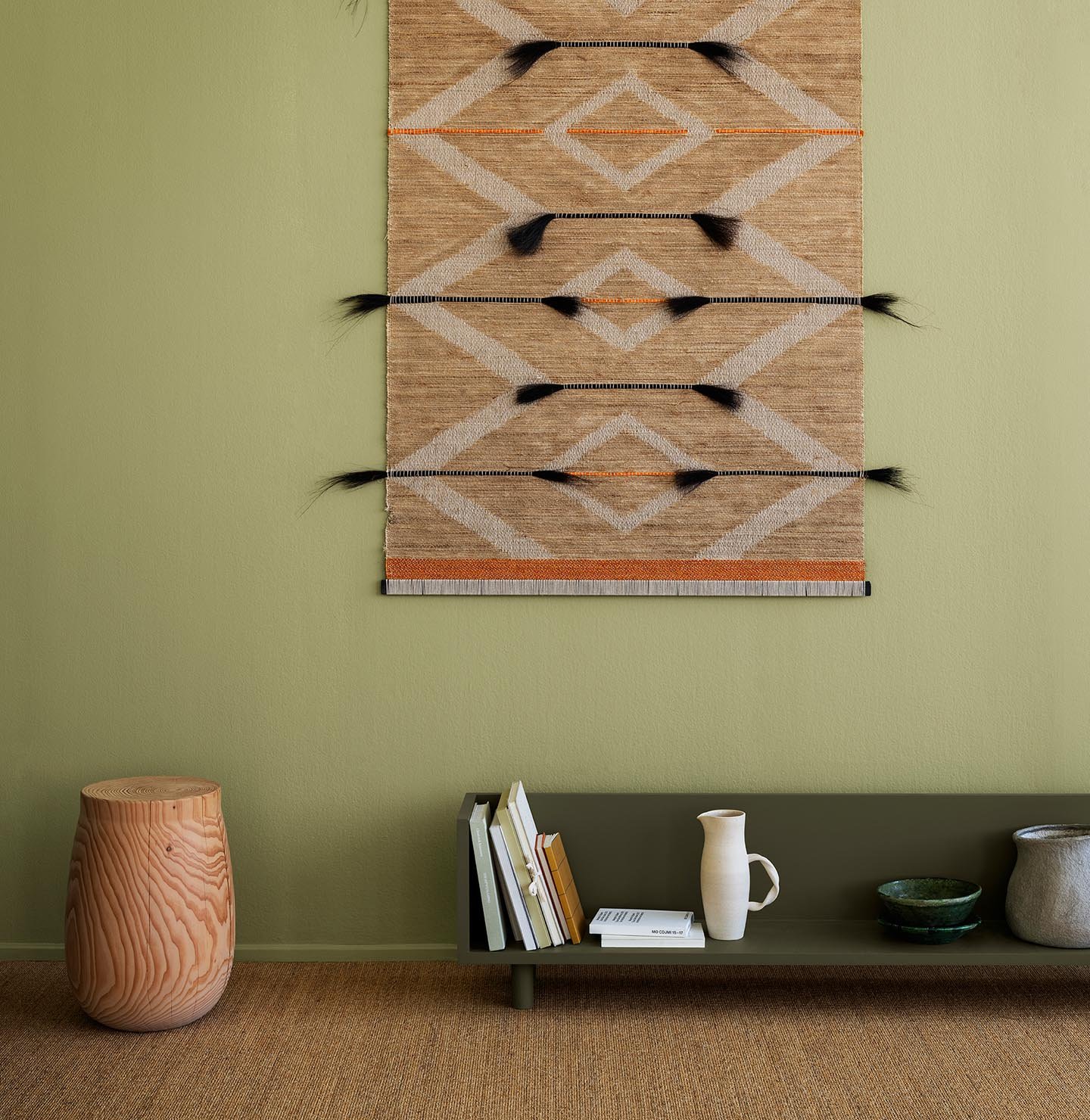 Böcker och keramik vilar på en grön hylla i färgen 8575 Natural Green från Jotun, med en vävd matta på väggen och en mjuk olivgrön bakgrund målad med Jotun 8183 Örtagård, som tillför naturlig värme till rummet.