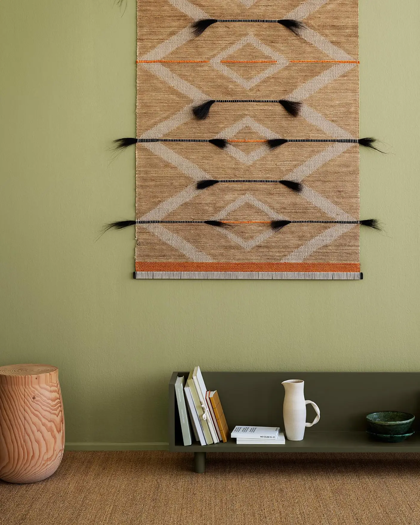 Böcker och keramik vilar på en grön hylla i färgen 8575 Natural Green från Jotun, med en vävd matta på väggen och en mjuk olivgrön bakgrund målad med Jotun 8183 Örtagård, som tillför naturlig värme till rummet.