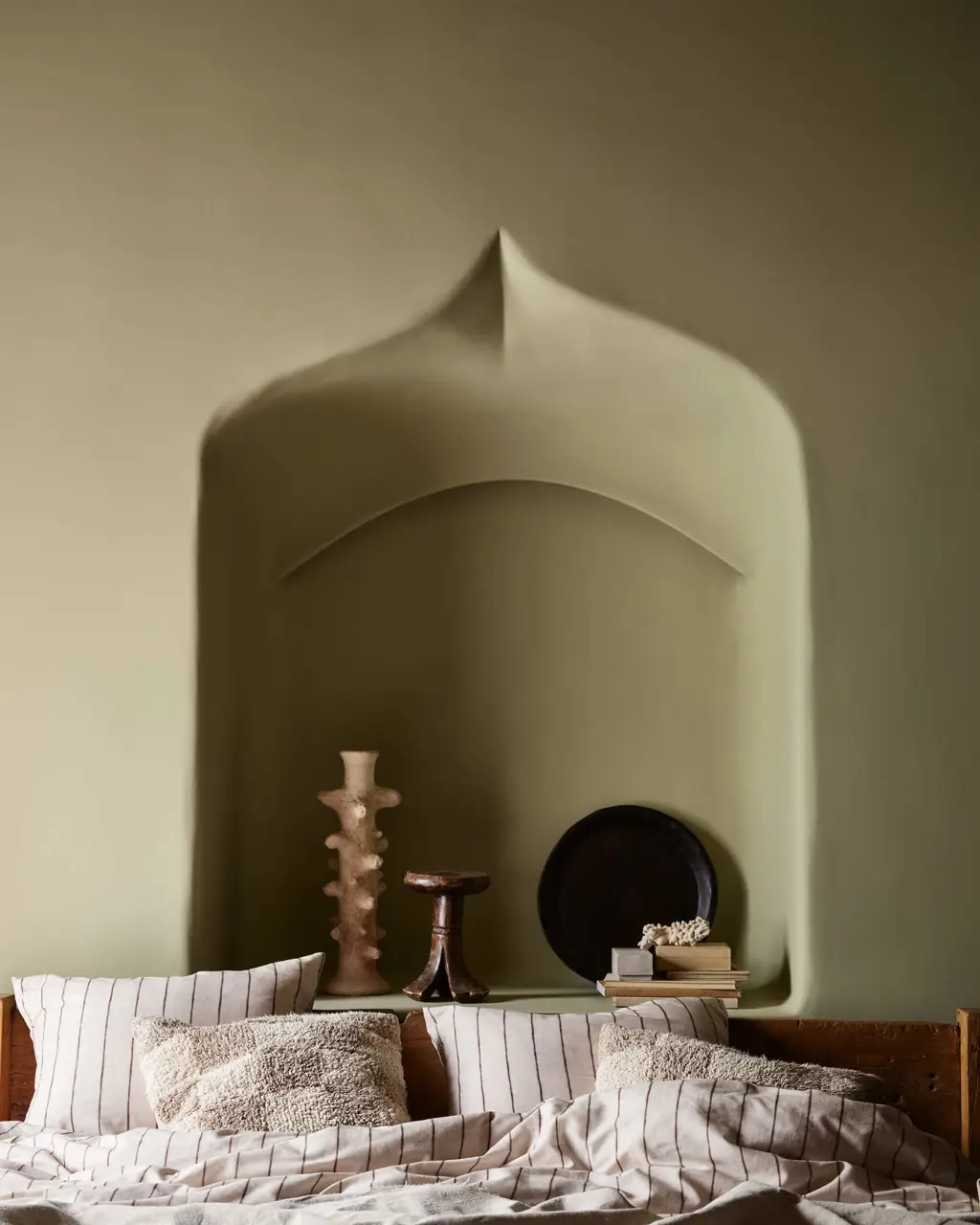 A cozy bedroom featuring olive green walls in Jotun 0290 Biri, a large sculpted arched alcove behind the bed, and striped linen bedding