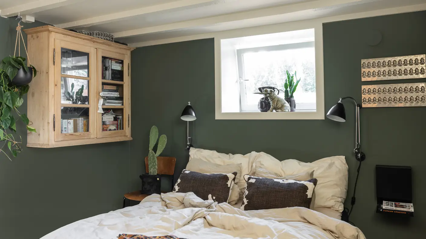 Soverom i underetasje med beige tak i Jotun Soft Dunes og mørke grønne vegger malt i Jotun Northern Mystic, industriell og personlig stil på interiøret og flere grønne planter.