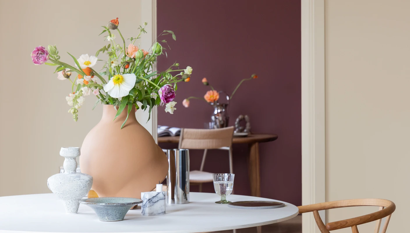 Et rundt spisebord med en organisk formet vase med fargerike, vårlige blomster, omgitt av den varme beigefargen JOTUN 1303 Observe, med et glimt inn i et kontor i varm rød farge malt i JOTUN 2951 Sophisticated Red. 