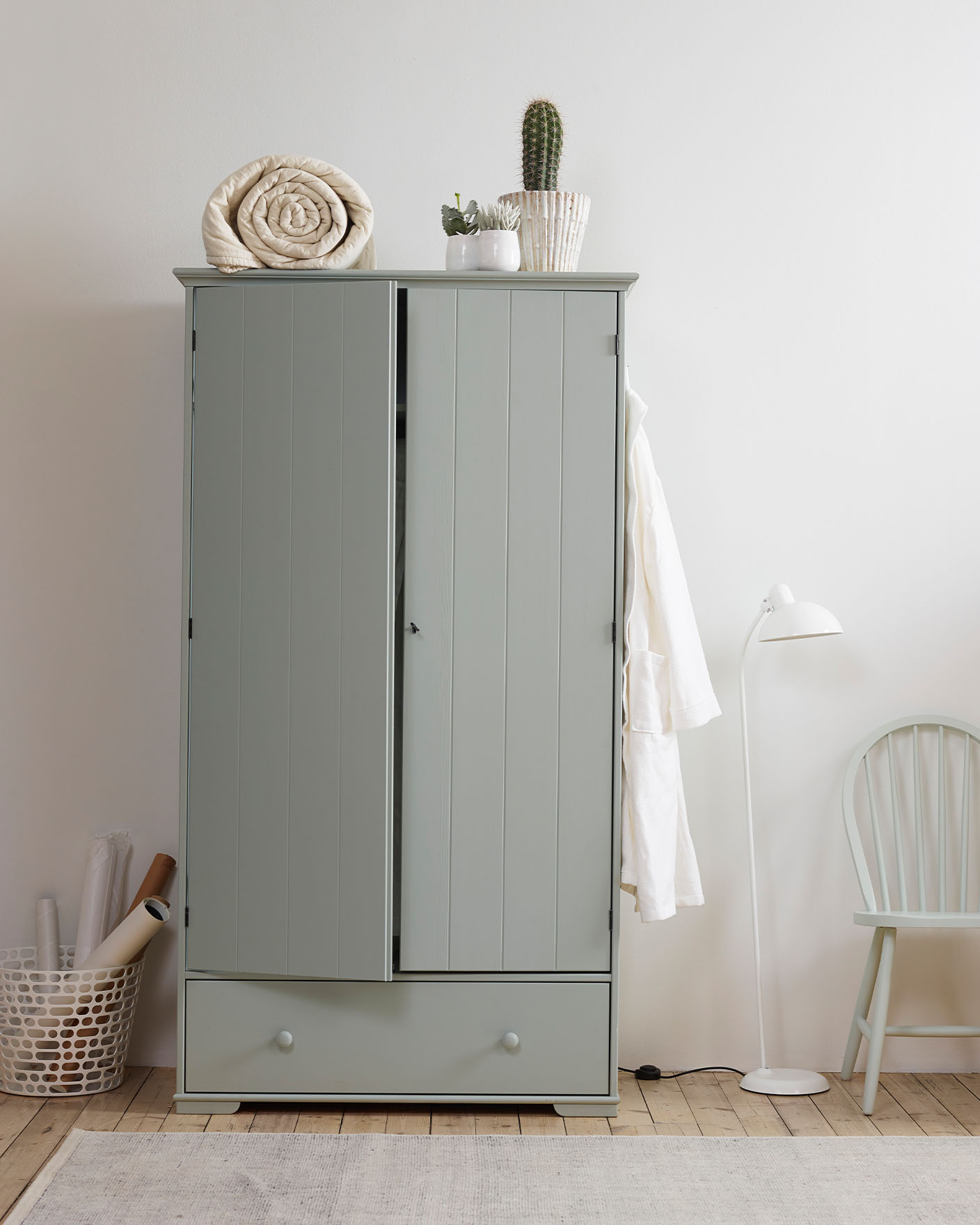 En dämpad, mintgrön garderob målad med Jotun 7163 Minty Breeze, toppad med ett ihoprullat täcke och krukväxter, bredvid en matchande stol och en vit golvlampa i ett lugnt, minimalistiskt sovrum.
