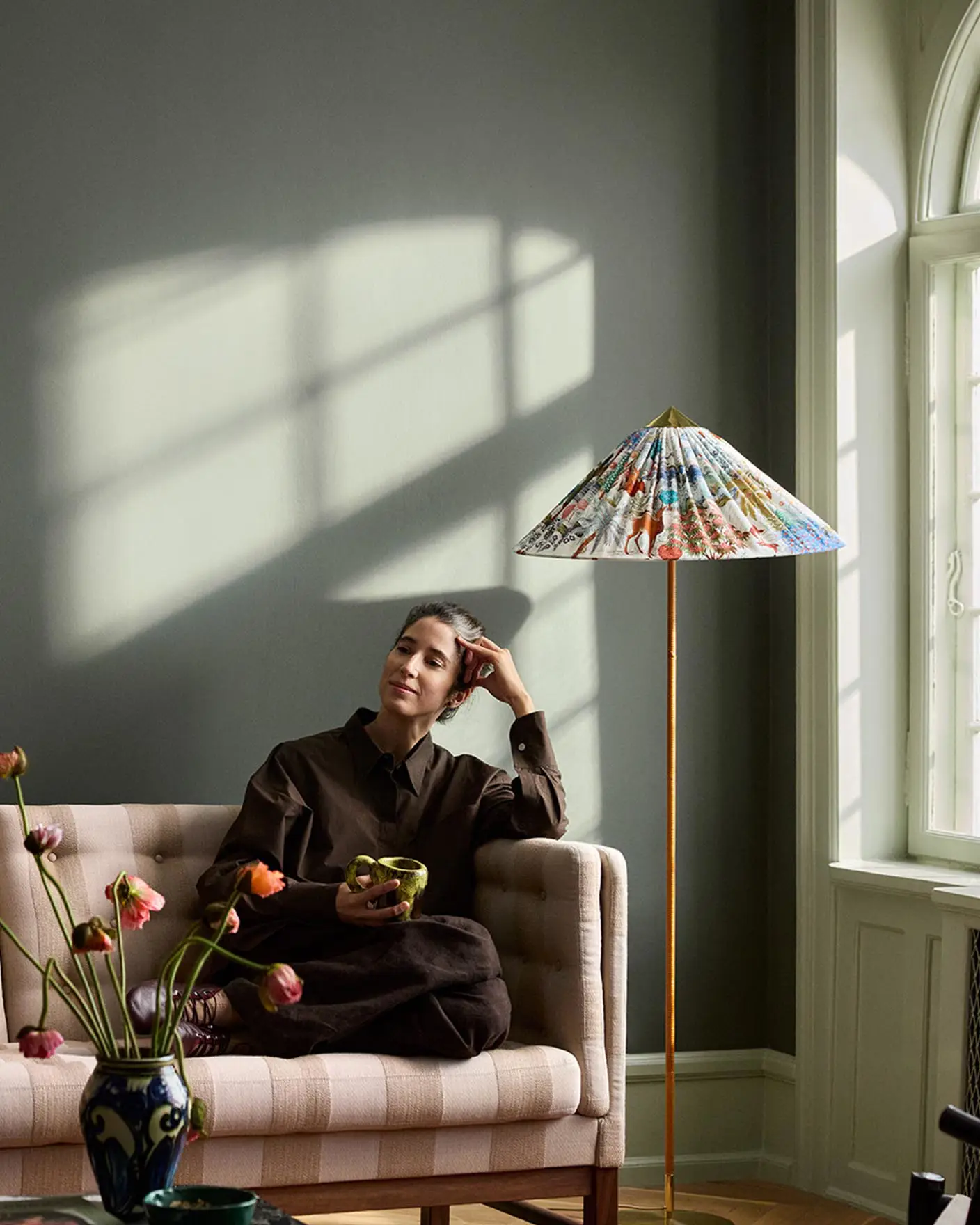 Woman sitting in a sofa with sunlight streaming through a window. The background wall is painted with the dark, muted green colour 7613 Northern Mystic from Jotun.