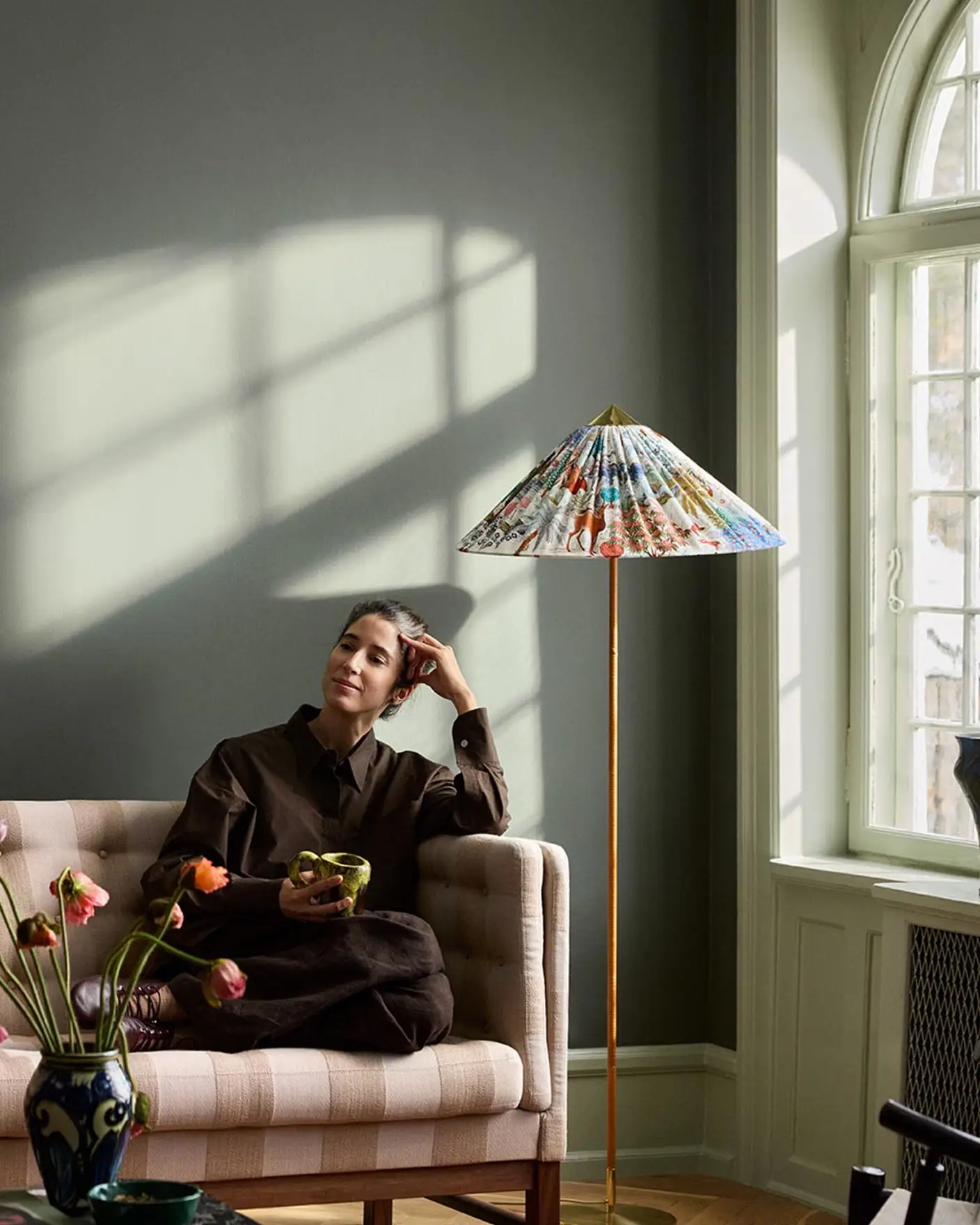 Woman sitting in a sofa with sunlight streaming through a window. The background wall is painted with the dark, muted green colour 7613 Northern Mystic from Jotun.