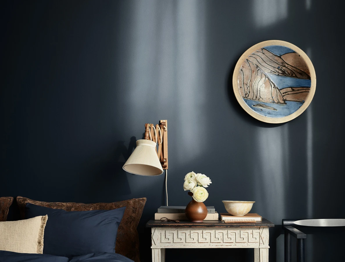 Et stemningsfullt soverom med dype, marineblå vegger i Jotun 4403 Midnight Blue, et vintage hvitt nattbord, en sakselampe på veggen og matchende blått sengetøy.
