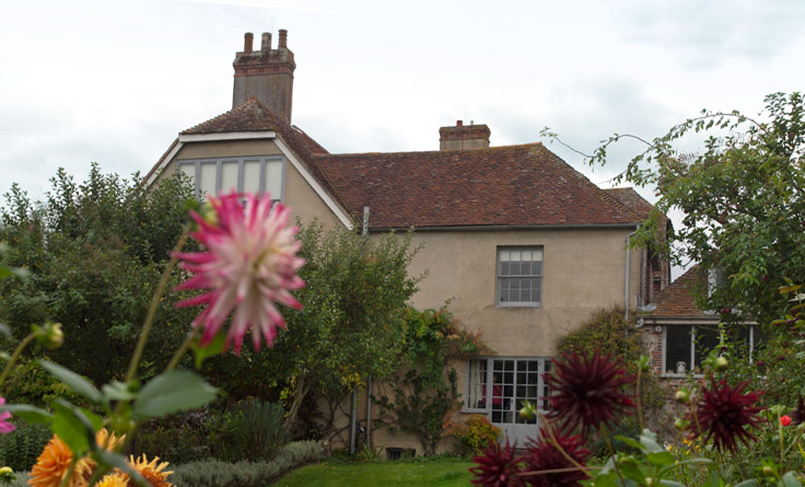 Historic English country house with red-tiled roof, surrounded by a lush garden and blooming flowers, under a cloudy sky