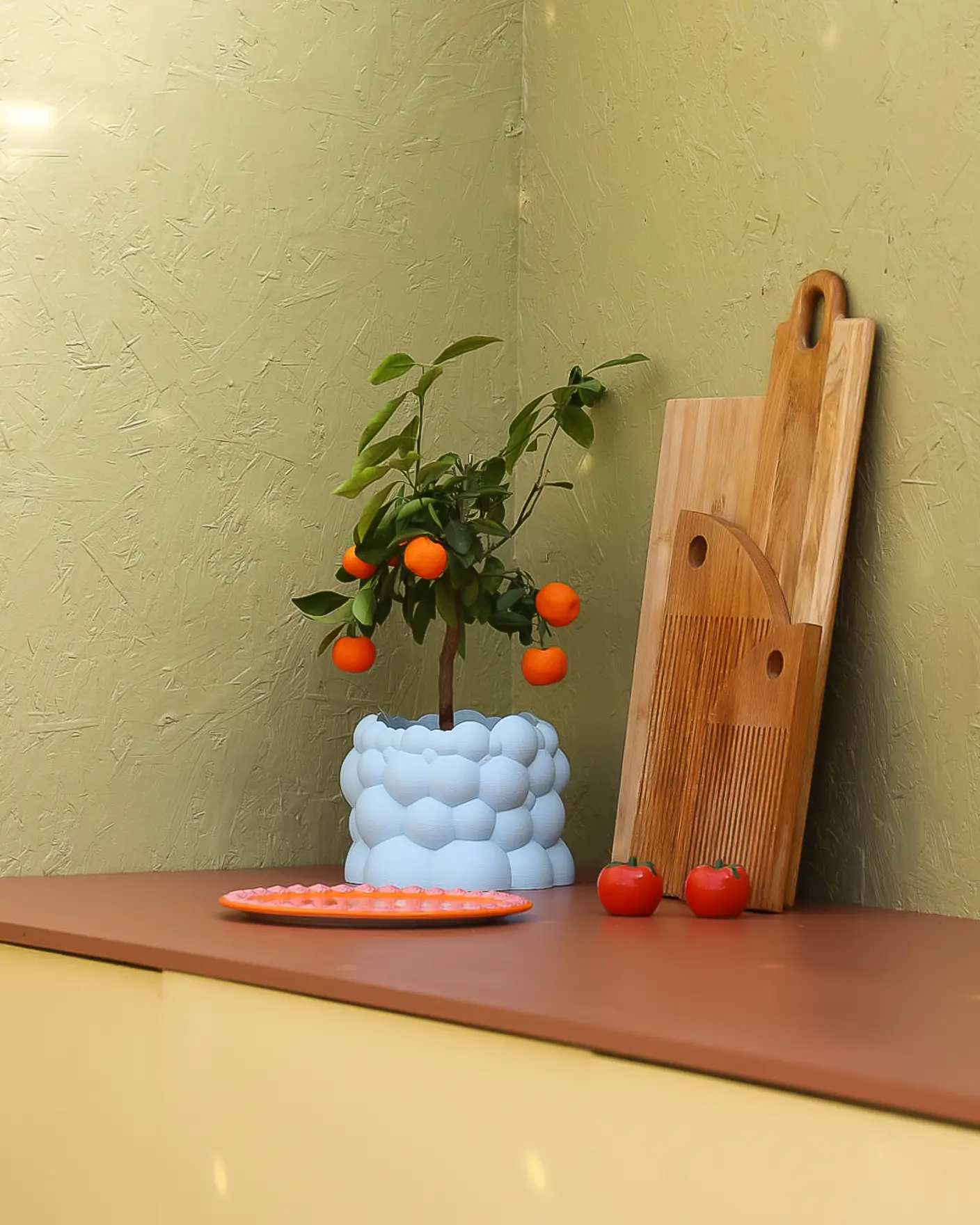 Detalj fra kjøkkenbenk med appelsinplante i lyseblå krukke og trefjøler, rød benkeplate i Jotun Amber Red, gult utekjøkken i Jotun Demigul og grønn vegg i Jotun Grønnland.