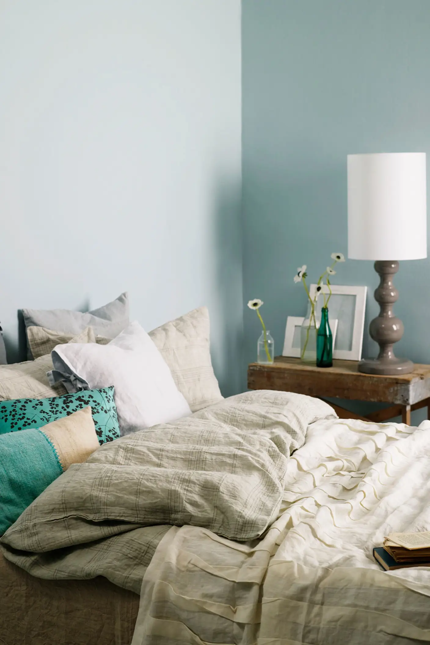 Inviting bedroom with layered neutral bedding, turquoise accent pillows, and a rustic wooden side table, set against muted, greenish blue wall painted with Jotun 5225 Lake View.