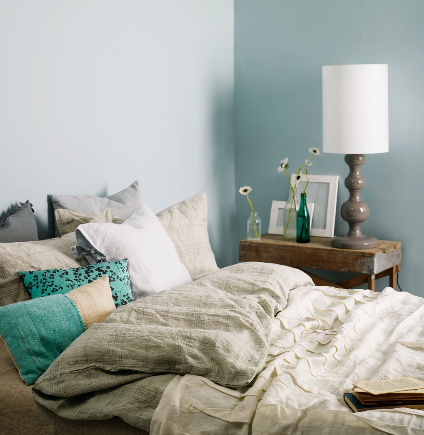 Inviting bedroom with layered neutral bedding, turquoise accent pillows, and a rustic wooden side table, set against muted, greenish blue wall painted with Jotun 5225 Lake View.