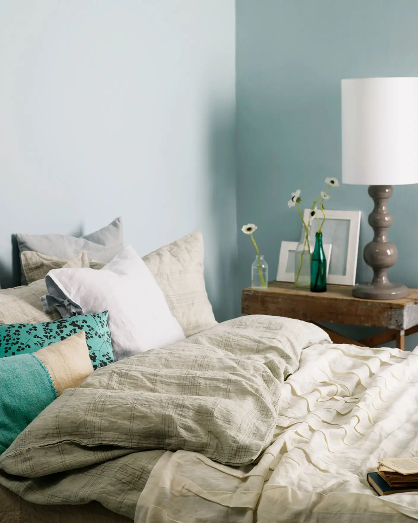 Inviting bedroom with layered neutral bedding, turquoise accent pillows, and a rustic wooden side table, set against muted, greenish blue wall painted with Jotun 5225 Lake View.