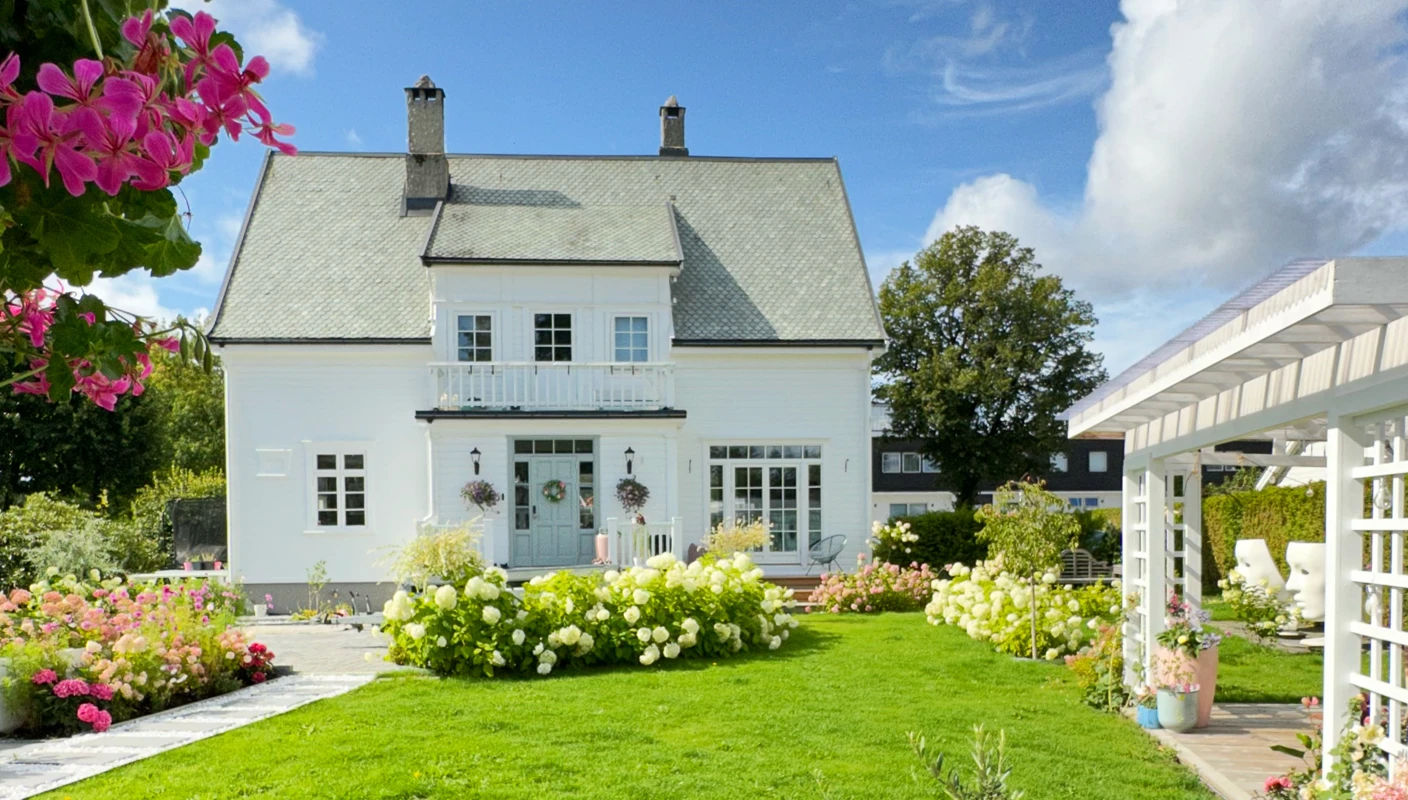 Hvitt trehus malt i Jotun 9918 Klassisk Hvit med DRYGOLIN Power Clean, grønn og blomstrende hage i front, grønt gress og bed.