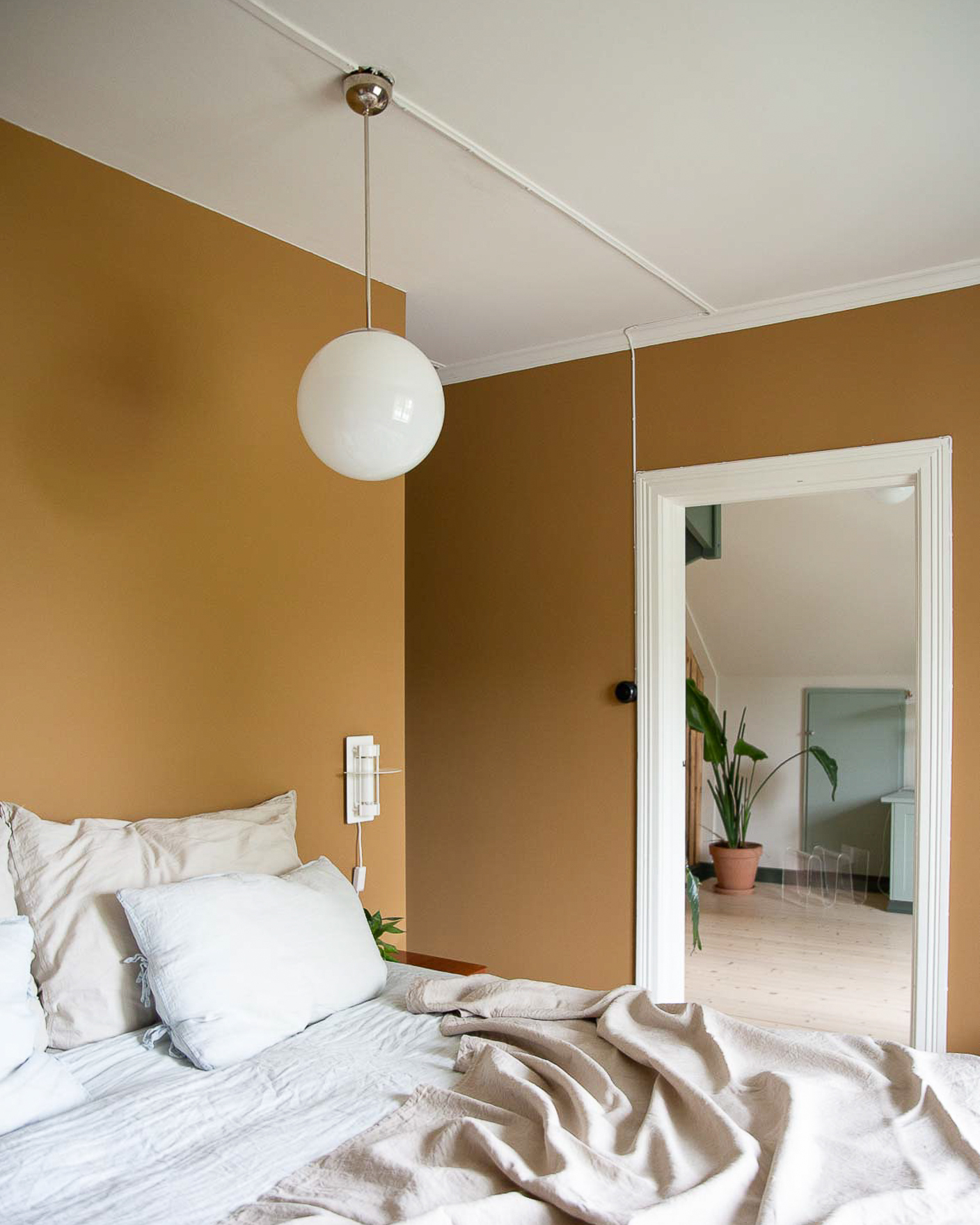 Soverom malt i mørkegule Jotun Dijon Yellow, hvitt tak og listverk i Jotun Bomull, seng med linfarget sengetøy og pendel i opalglass i tak.