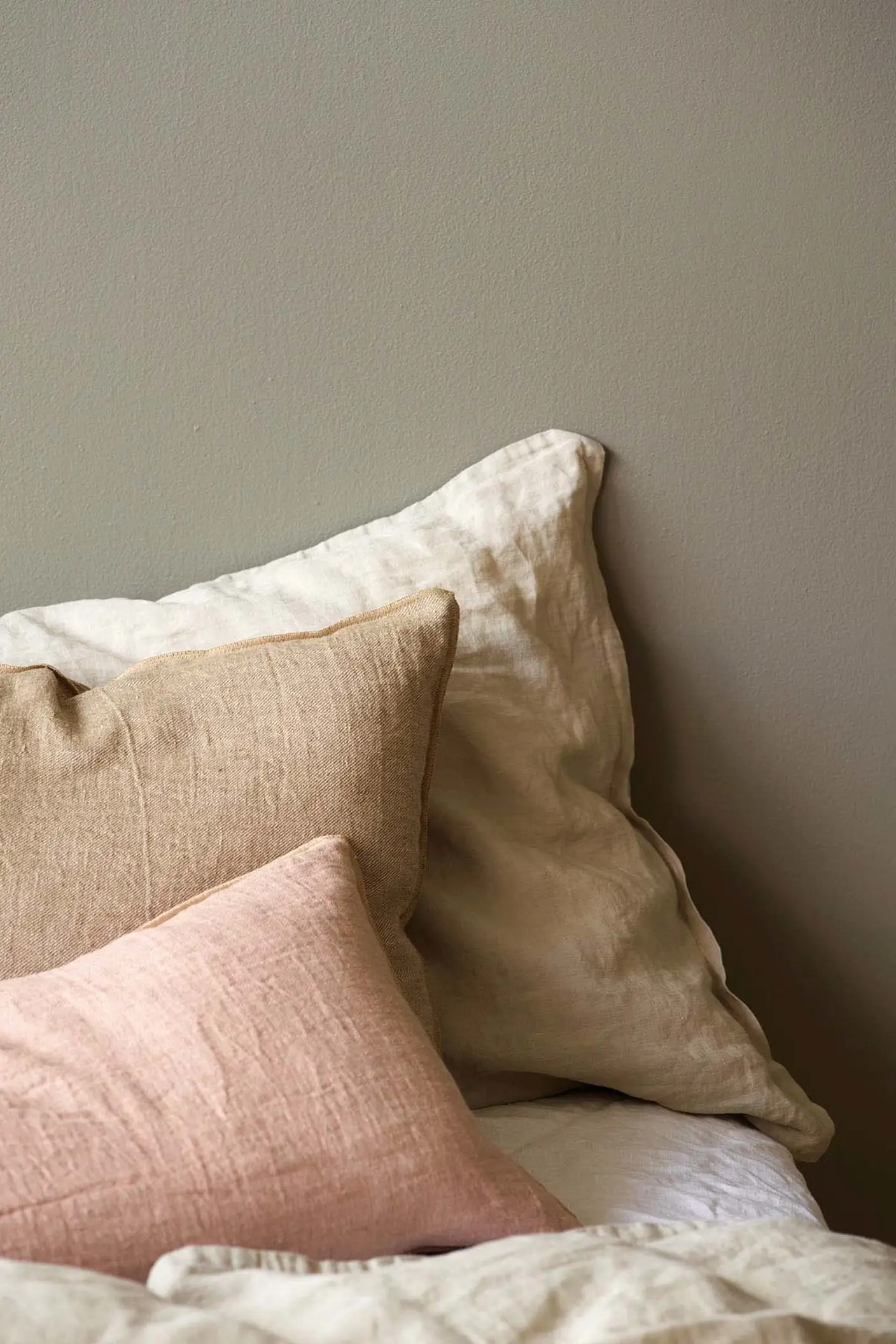 Bej, taba ve pembeye yakın tonlardaki keten yastıklarla hazırlanmış yatak, Jotun 1877 Pebblestone ile boyanmış altın gri bir duvarın önünde yer alıyor.