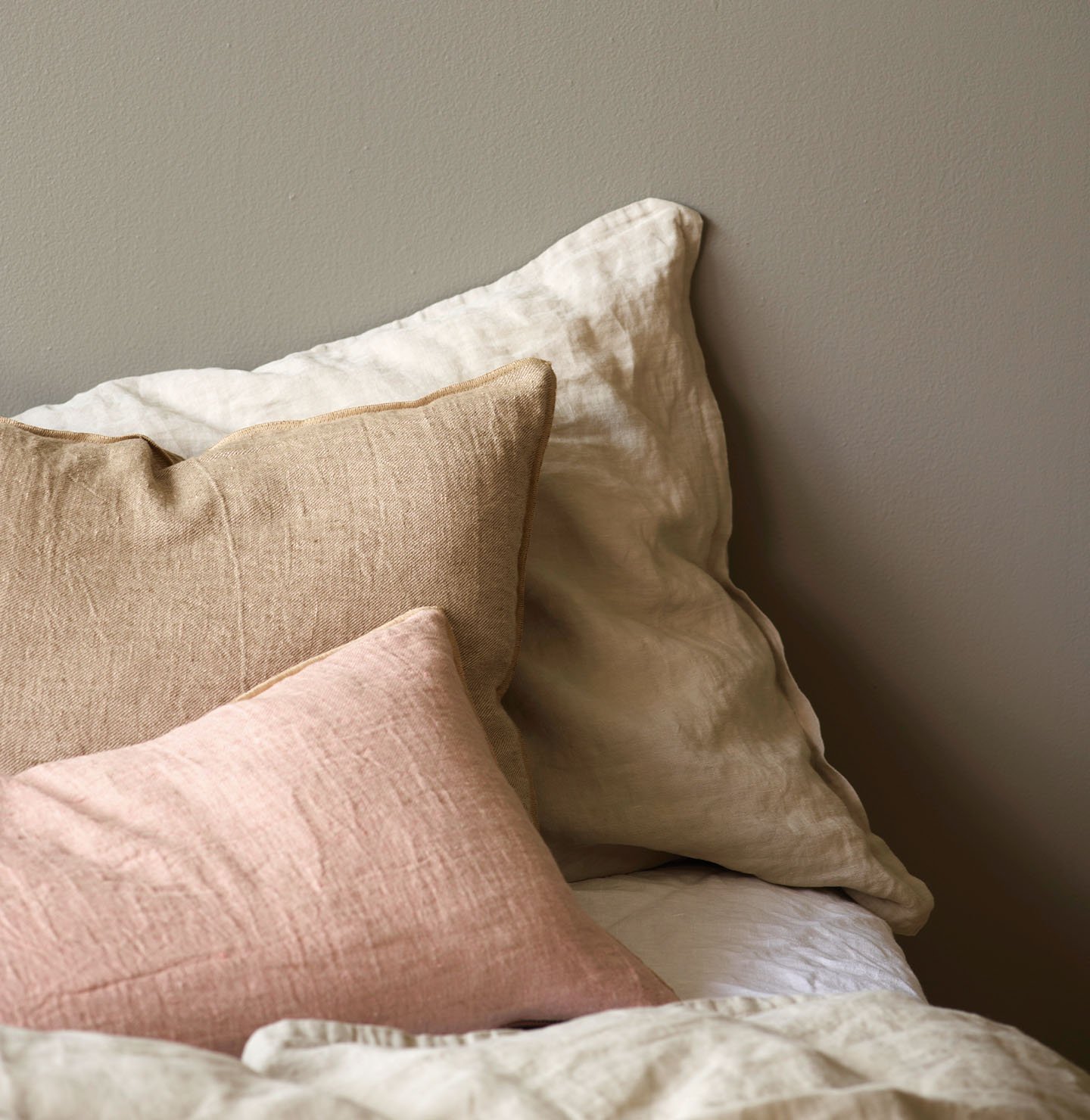 Close-up of a bed with linen pillows in soft beige, warm tan, and muted pink tones, set against a golden grey wall painted with Jotun 1877 Pebblestone.