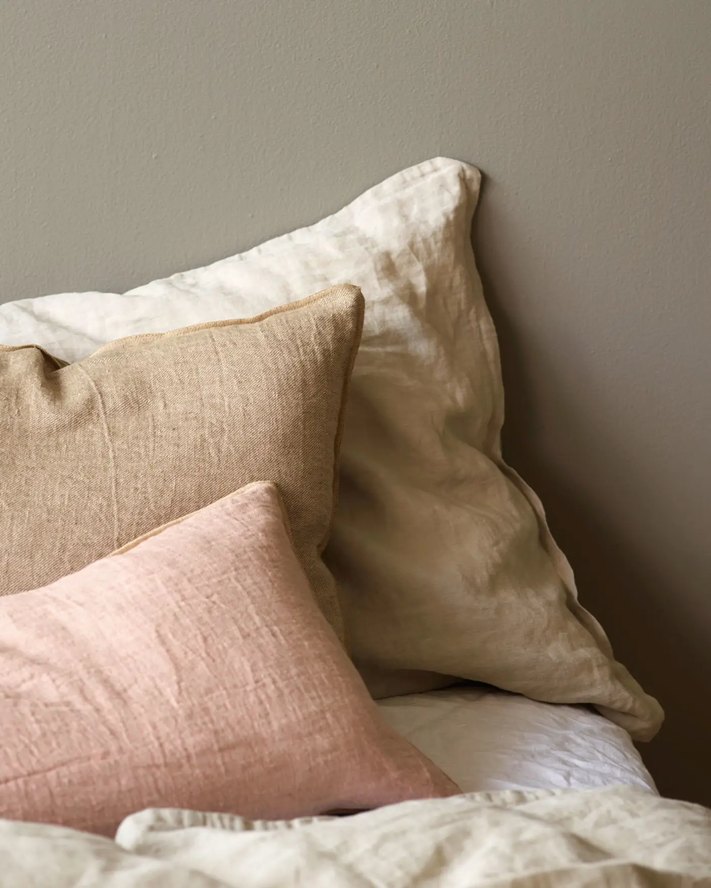 Close-up of a bed with linen pillows in soft beige, warm tan, and muted pink tones, set against a golden grey wall painted with Jotun 1877 Pebblestone.