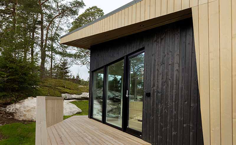 Moderne hytte med svart og naturlig trepanel, beiset med  TREBITT Matt Oljebeis i fargene Funkissort og Naturlig Eldet. Hytta er  omgitt av skog, sett fra treterrasse beiset lys grå med TREBITT Terrassebeis i fargen Shimmergrå, i en naturskjønn setting