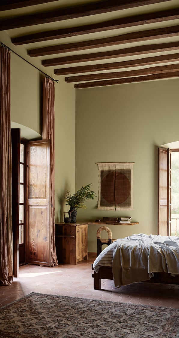 Rustic bedroom with exposed wood ceiling beams, open wooden doors, walls painted in the colour Jotun Biri, and a bed with linen bedding.