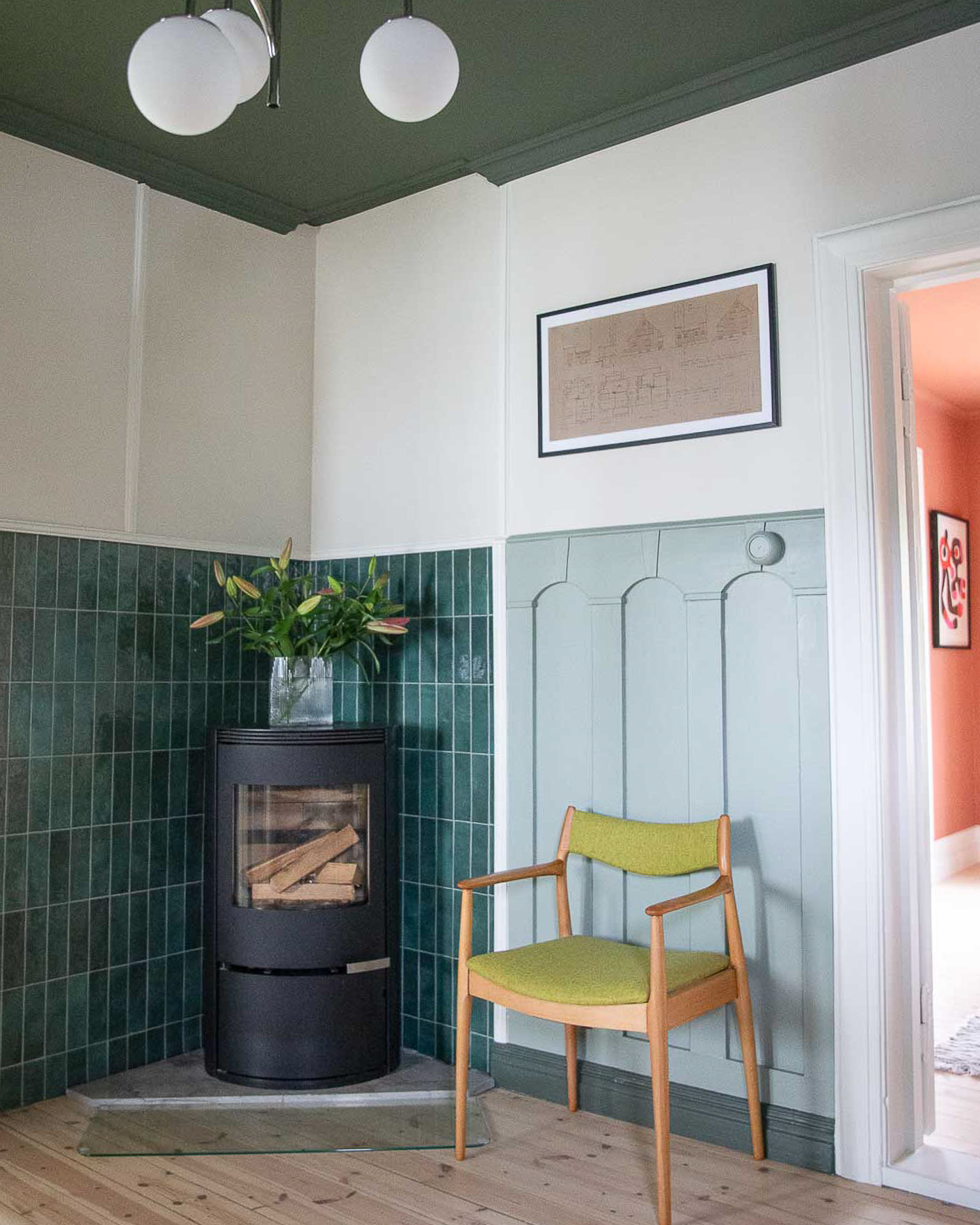 Hall i klassisk 1920-talls villa med vedovn med grønne fliser bak, brystningspanel malt lysegrønt med Jotun Treasure, vegg over panel malt beige med Jotun Sans og tak og fotlist malt mørkegrønt i Jotun Northern Mystic.