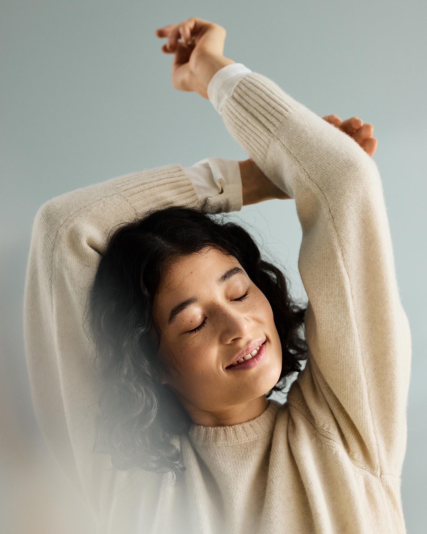 Woman with curly hair with arms raised above the head, in front of a muted blue background painted with the colour 6378 Iconic from Jotun.