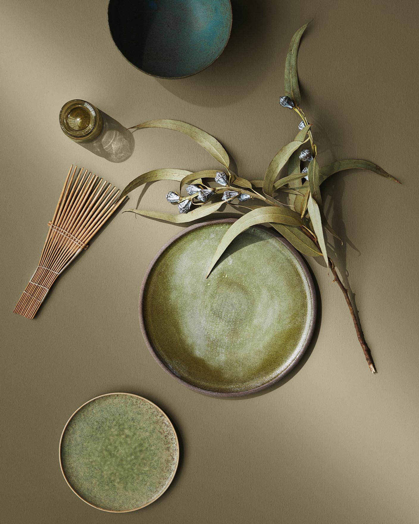 Green-toned ceramics and natural elements lie on a warm olivengreen surface painted with the colour 8546 Local Green from Jotun, creating a soft and earthy composition.