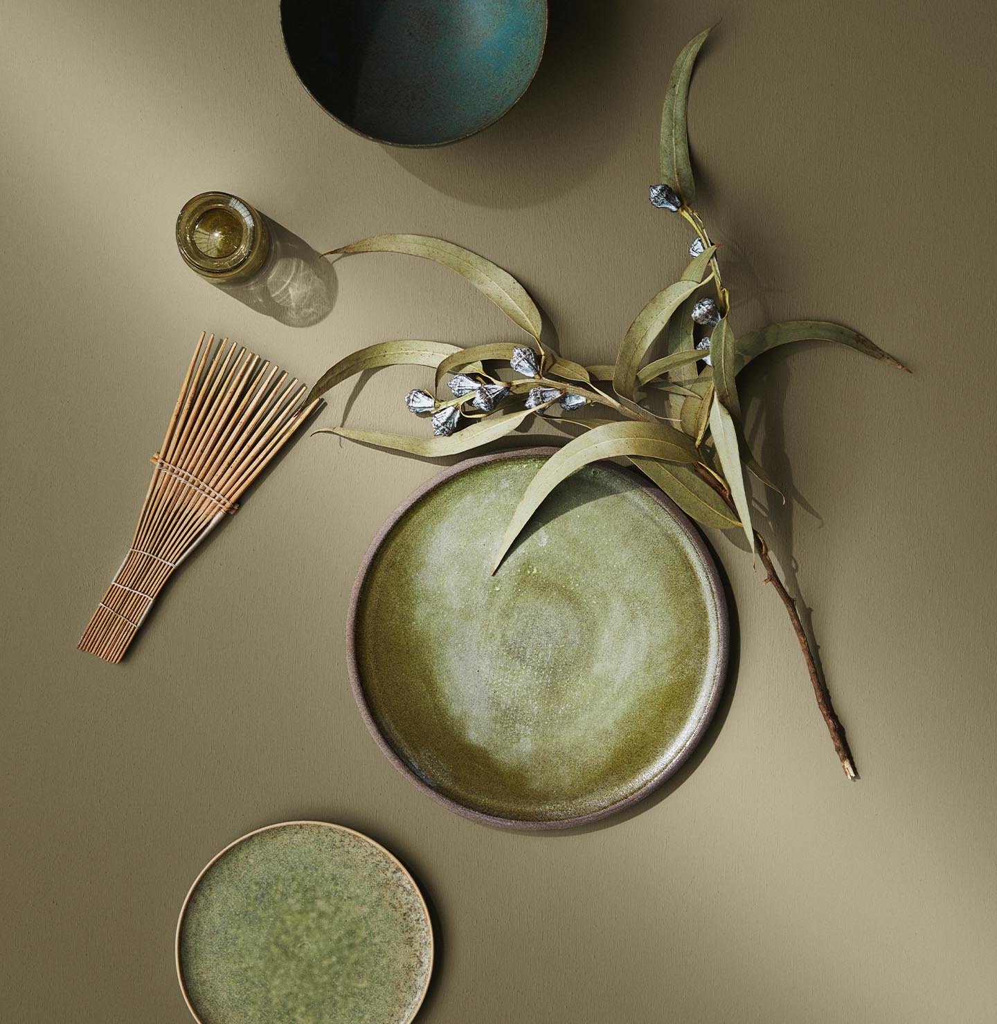 Green-toned ceramics and natural elements lie on a warm olivengreen surface painted with the colour 8546 Local Green from Jotun, creating a soft and earthy composition.