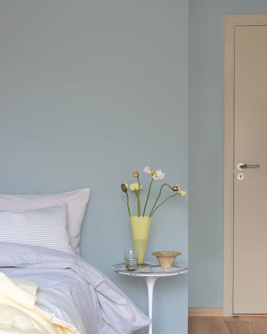 Sovrum med fräscha linnetextilier och gula blommor på nattduksbordet. Väggarna är målade i den blågröna kulören JOTUN 6378 Iconic och dörren med listverk i den mörkbeige tonen JOTUN 12300 Hazelnut Beige.